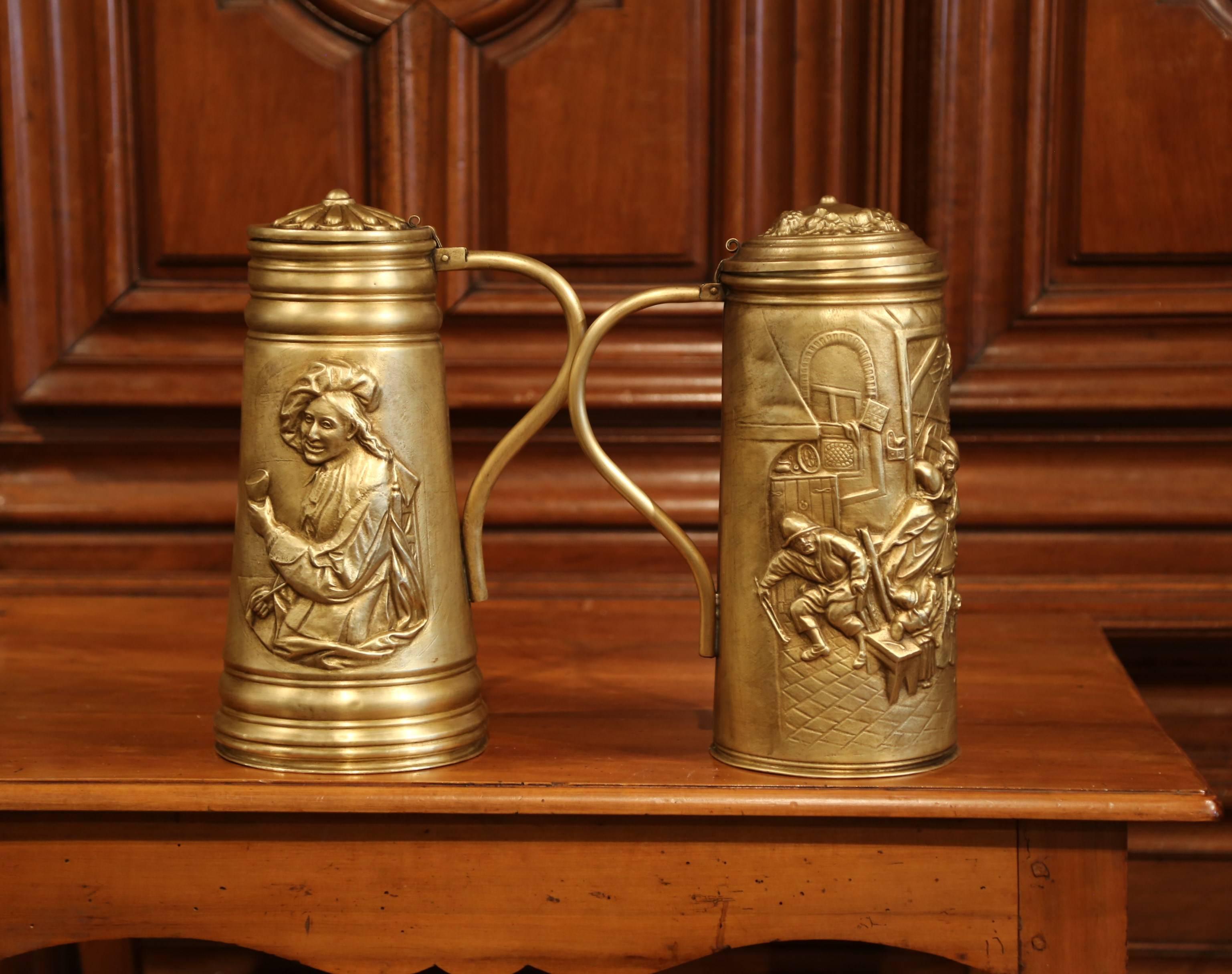 Decorate your bar with this pair of antique brass beer mugs, crafted in Belgium circa 1880, the pitchers with handles and lids, feature interesting repousse work including a tavern scene in the manner of Teniers on one. The other pitcher features a