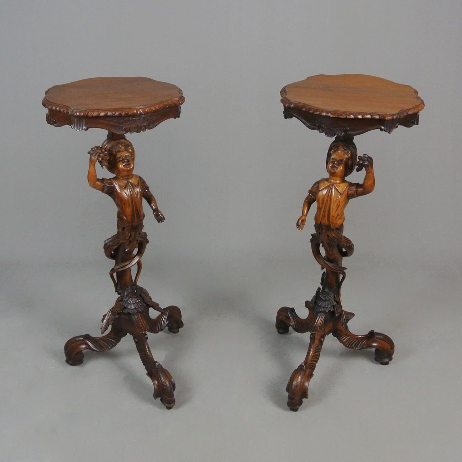 A wonderful and rare pair of mid 19th century solid walnut Black Forest torcheres, each column formed as a well carved boy, carrying a bunch of grapes in one hand and wearing a smock shirt and cap.  Their bodies transform into a tree trunk wrapped