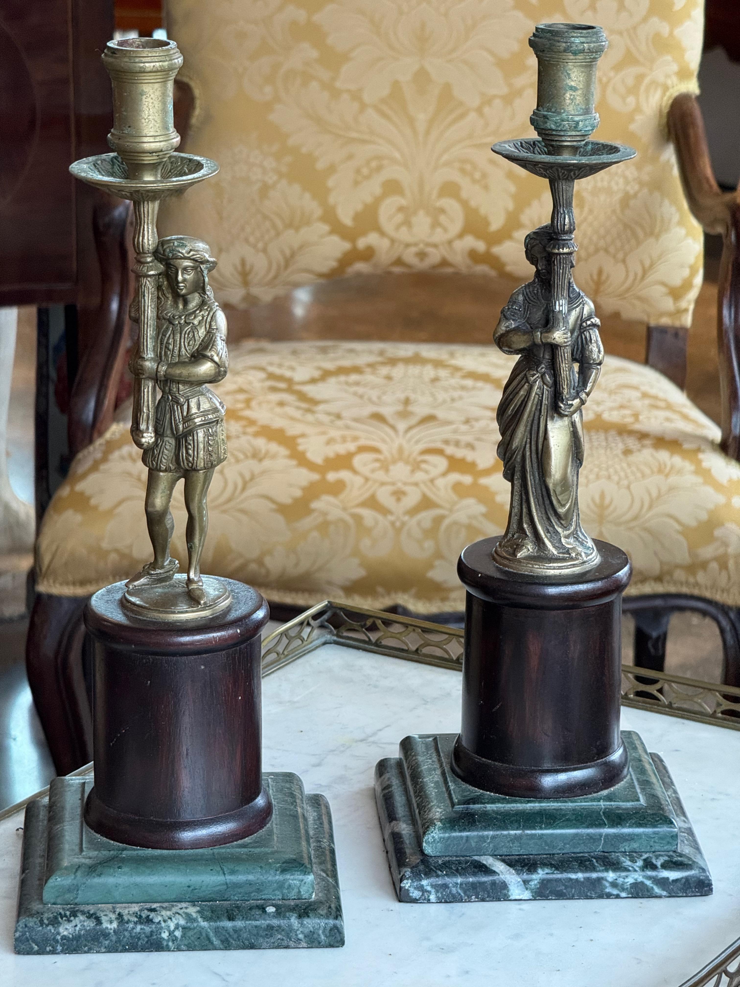 Coppia di candelieri in ottone fuso finemente dettagliati che raffigurano figure classiche maschili e femminili, ciascuno montato su una base in marmo verde e legno. Le figure sono rese in modo nitido e aggiungono un tocco decorativo alle basi