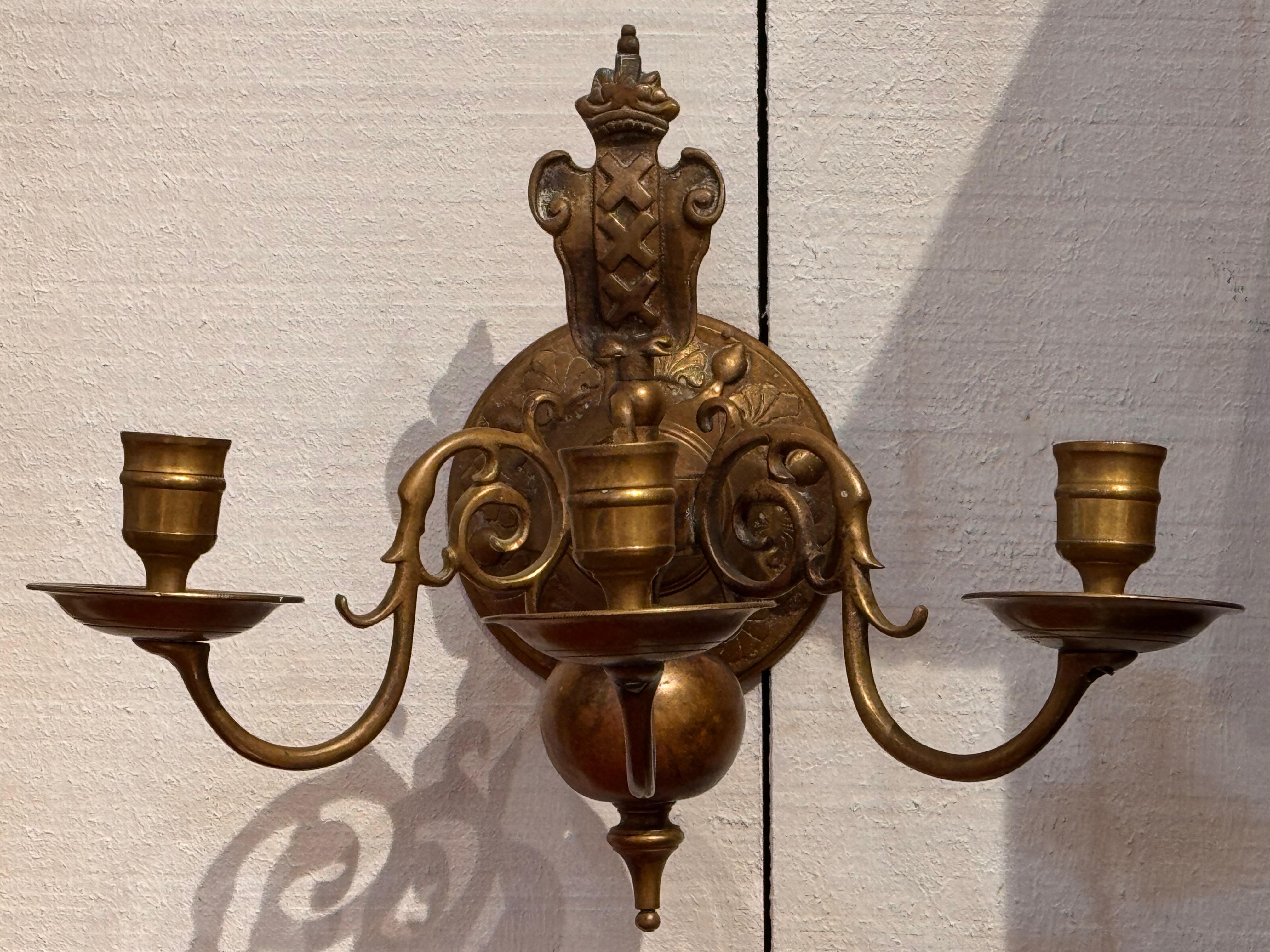 This pair of 19th century brass sconces features three scrolling candle arms anchored by a baluster-form body and decorative backplate with stylized shell and foliate motifs. The upper finial is shaped with classical detail, adding architectural