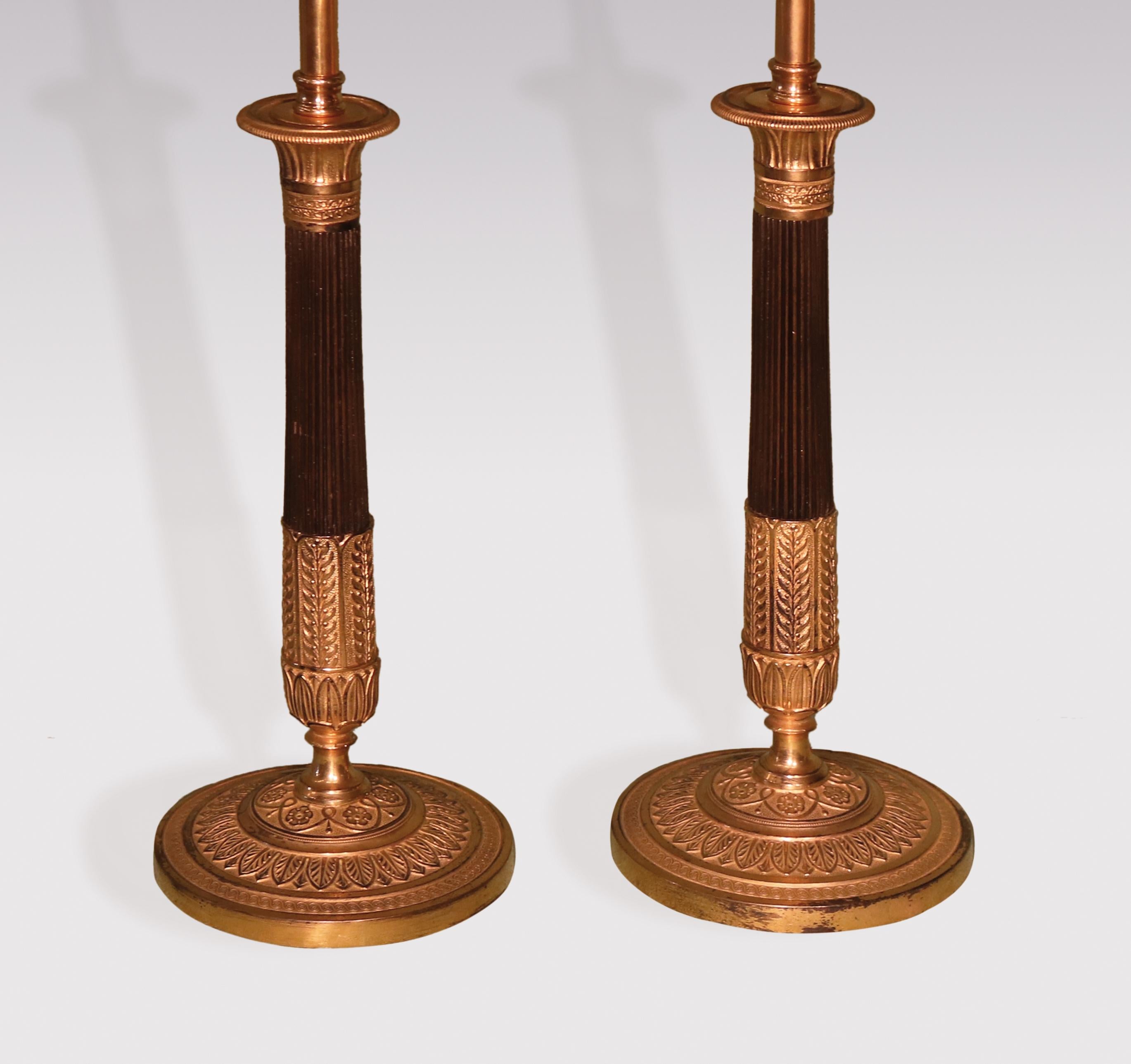 A pair of early 19th century bronze & ormolu candlesticks, having lotus leaf sconces above reeded tapering stems with leaf & berry decoration, raised on leaf & flower decorated circular bases. (Now converted to lamps), height of candlesticks 10