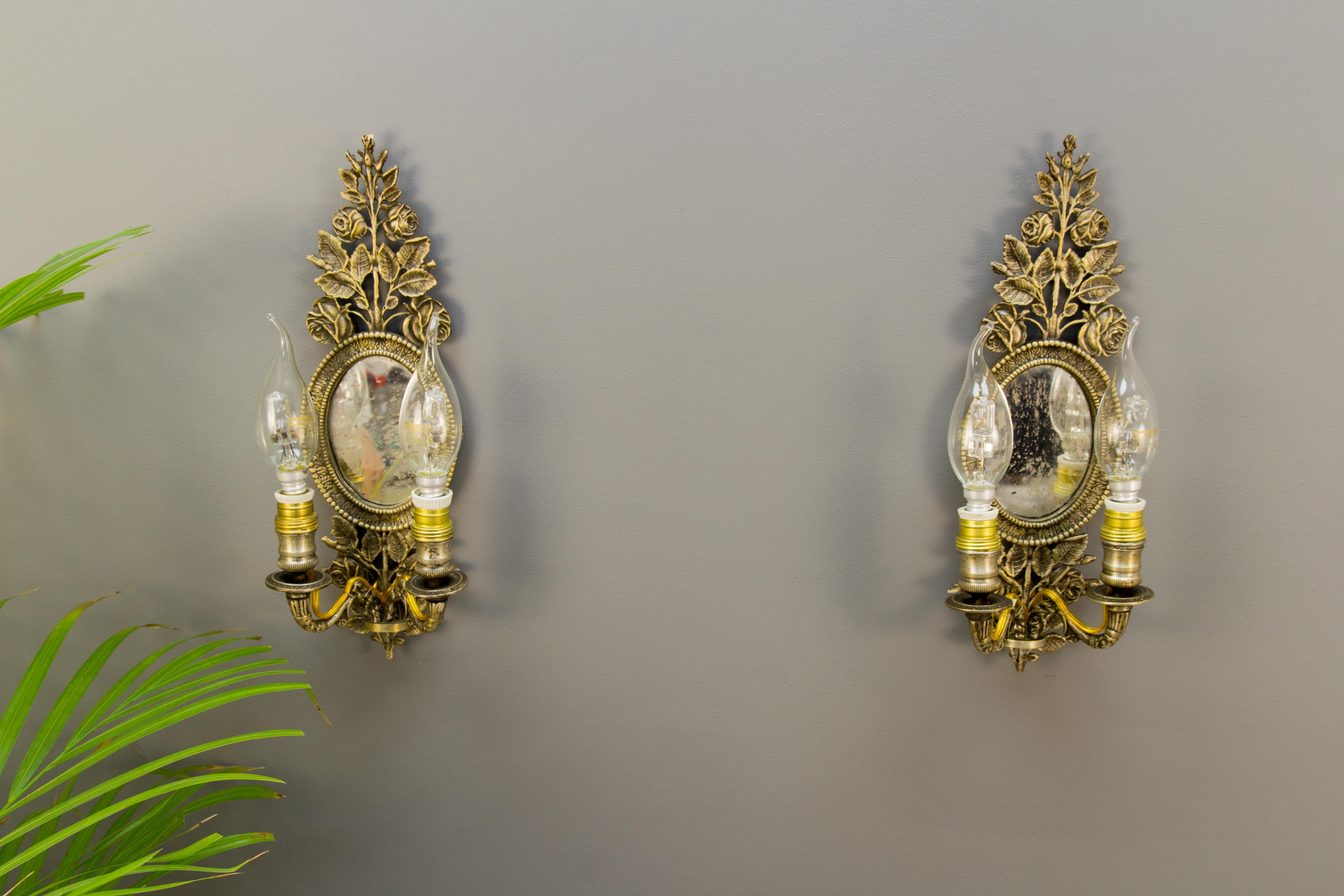 A pair of small but very fine antique French wall sconces with oval framed mirrors and beautiful floral details, roses. Originally crafted for two candles each, these antique bronze wall lights have been previously electrified, now each has two