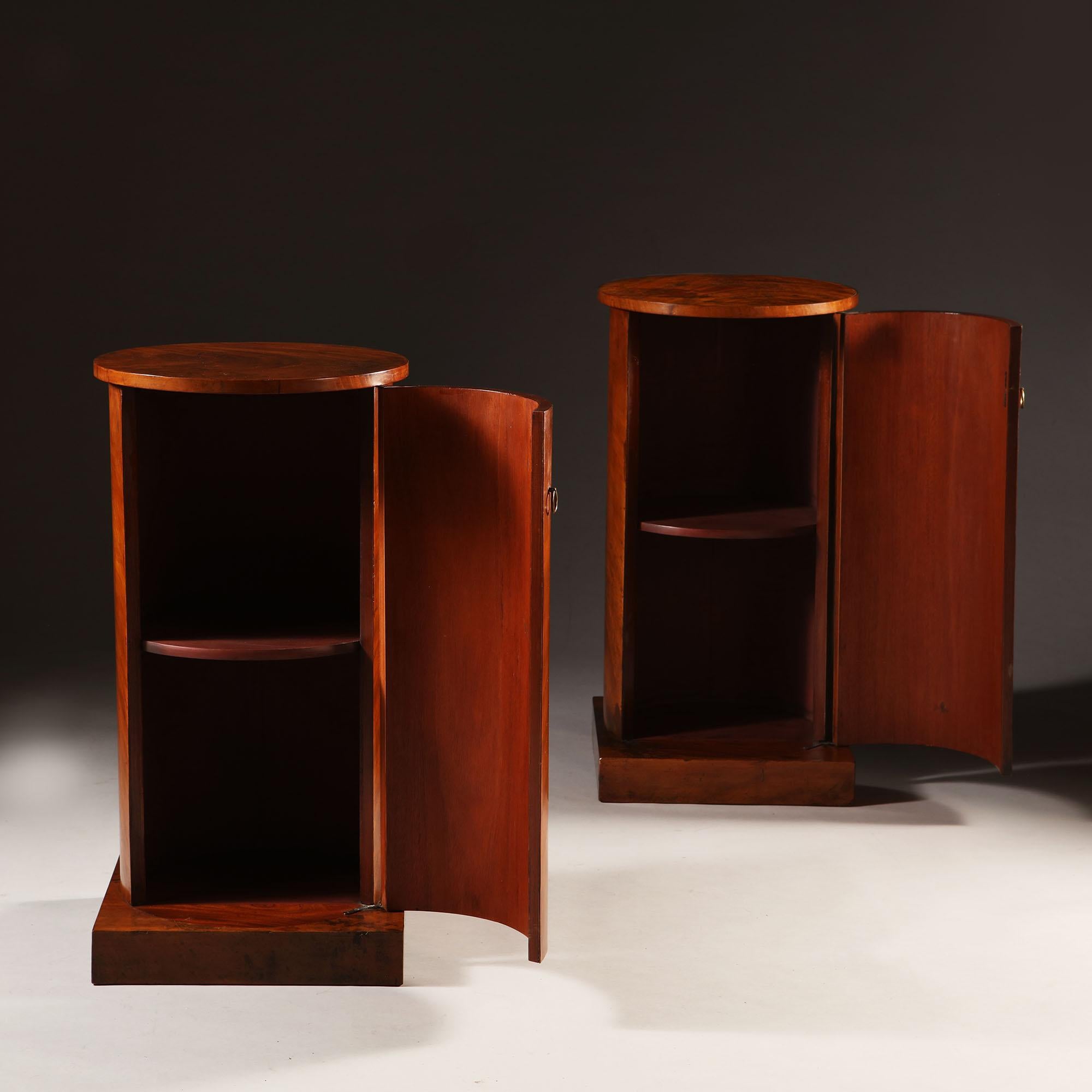 A pair of 19th century English flame mahogany wood bedside cupboards / tables in the form of pedestals, each with one door to the front opening to reveal two shelves, with brass loop handles to the doors, all supported on square plinth bases.