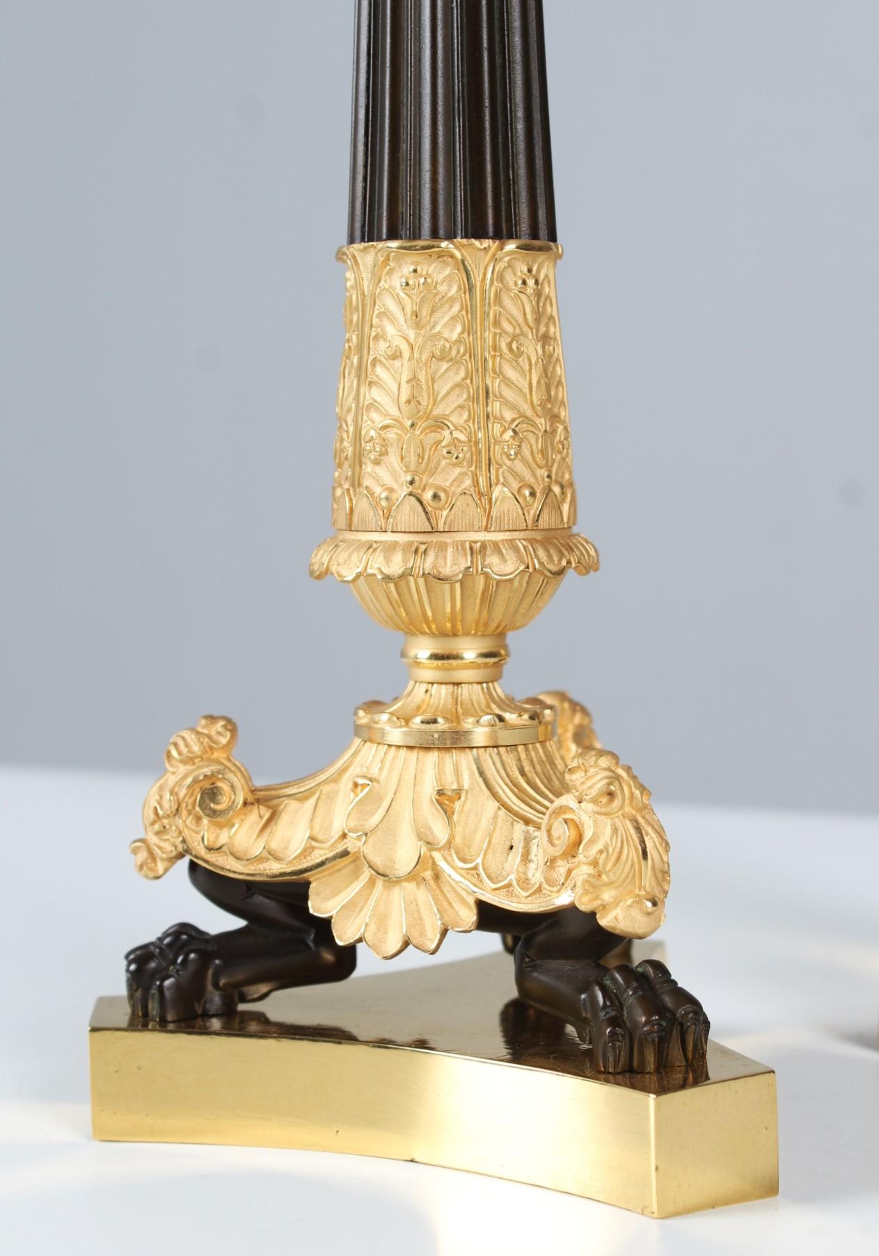 Pair of antique candlesticks. 

H x W x D: 37 x 13 x 13 cm.

On the triangular laterally indented plinth stand on each of three lion paws the partly gilded, partly patinated candlesticks. 
The lower part of the stem is decorated with acanthus