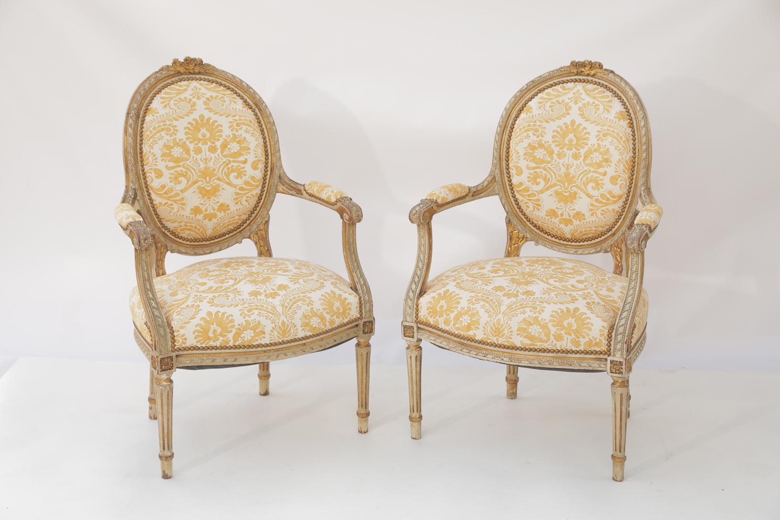 Pair of fauteuils in the Louis XVI style, having a painted and parcel-gilt finish showing natural wear, each oval padded backrest in a gadrooned frame, surmounted by foliate carving, outswept arms, with elbow rests, ending in scrolls, set on curved