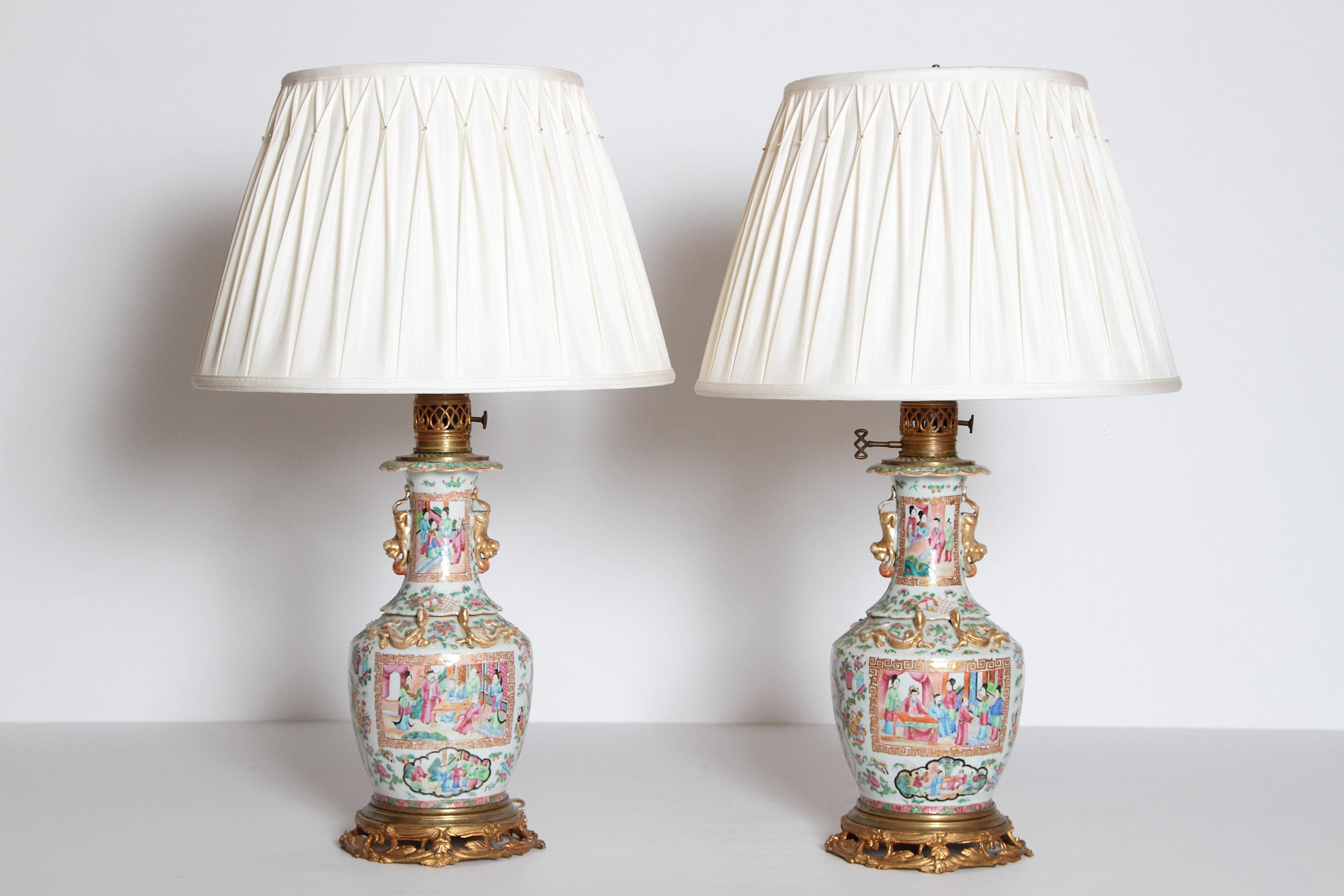 An elegant pair of Chinese rose medallion vases, originally custom mounted as oil lamps, now electrified, set on ormolu bases in foliate design. The bulbous shape of the vase contains two large panels of people bordered in a fret key motif and four
