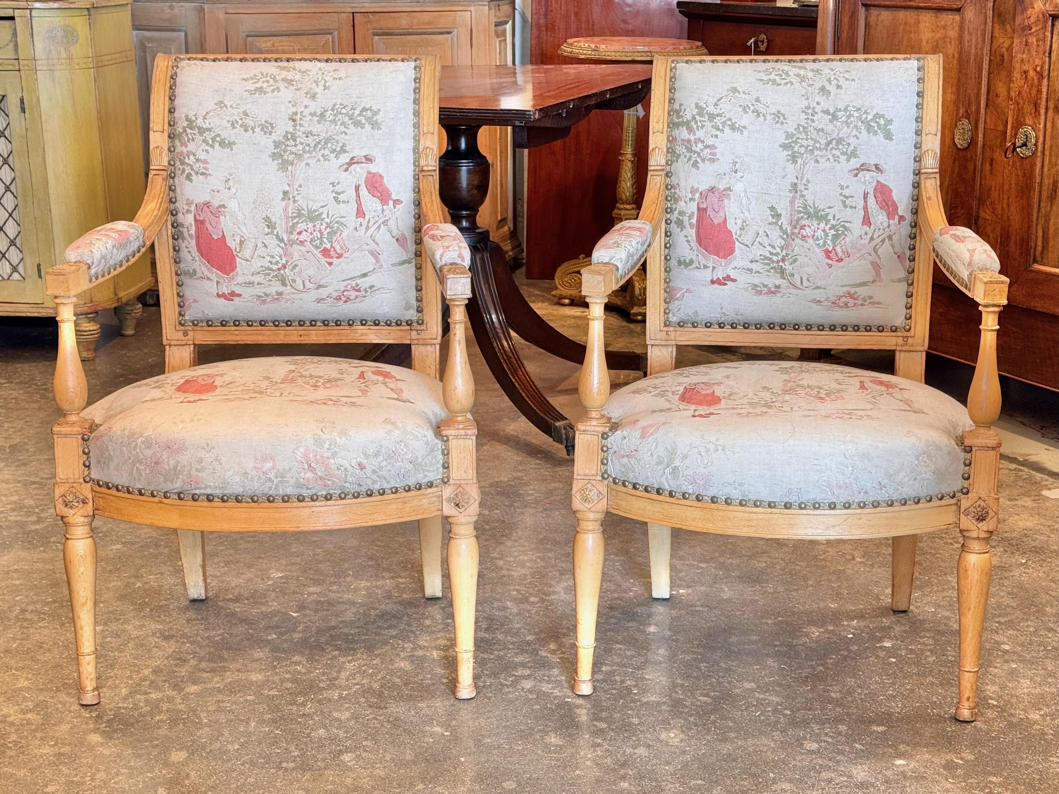 Beechwood fauteuils with classic Directoire lines, featuring turned and fluted legs, gracefully curved arms, and detailed joinery. The upholstery showcases a pastoral toile pattern with soft red and green tones, secured with brass nailhead trim. The