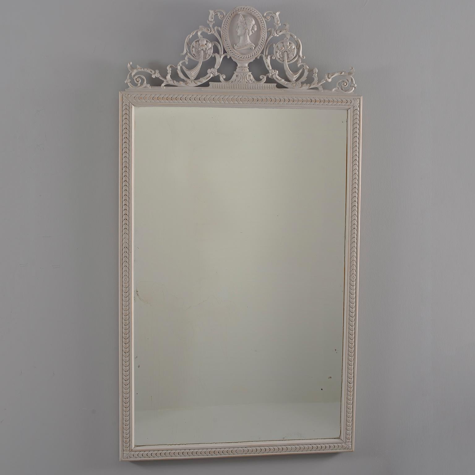 Pair of Directoire style wood framed mirrors with antique white painted finish feature beveled edges and elaborate crests topped with a carved bust. Sold and priced as a pair, circa 1890.  Actual Mirror Size:  36.75” h x 22.75” w