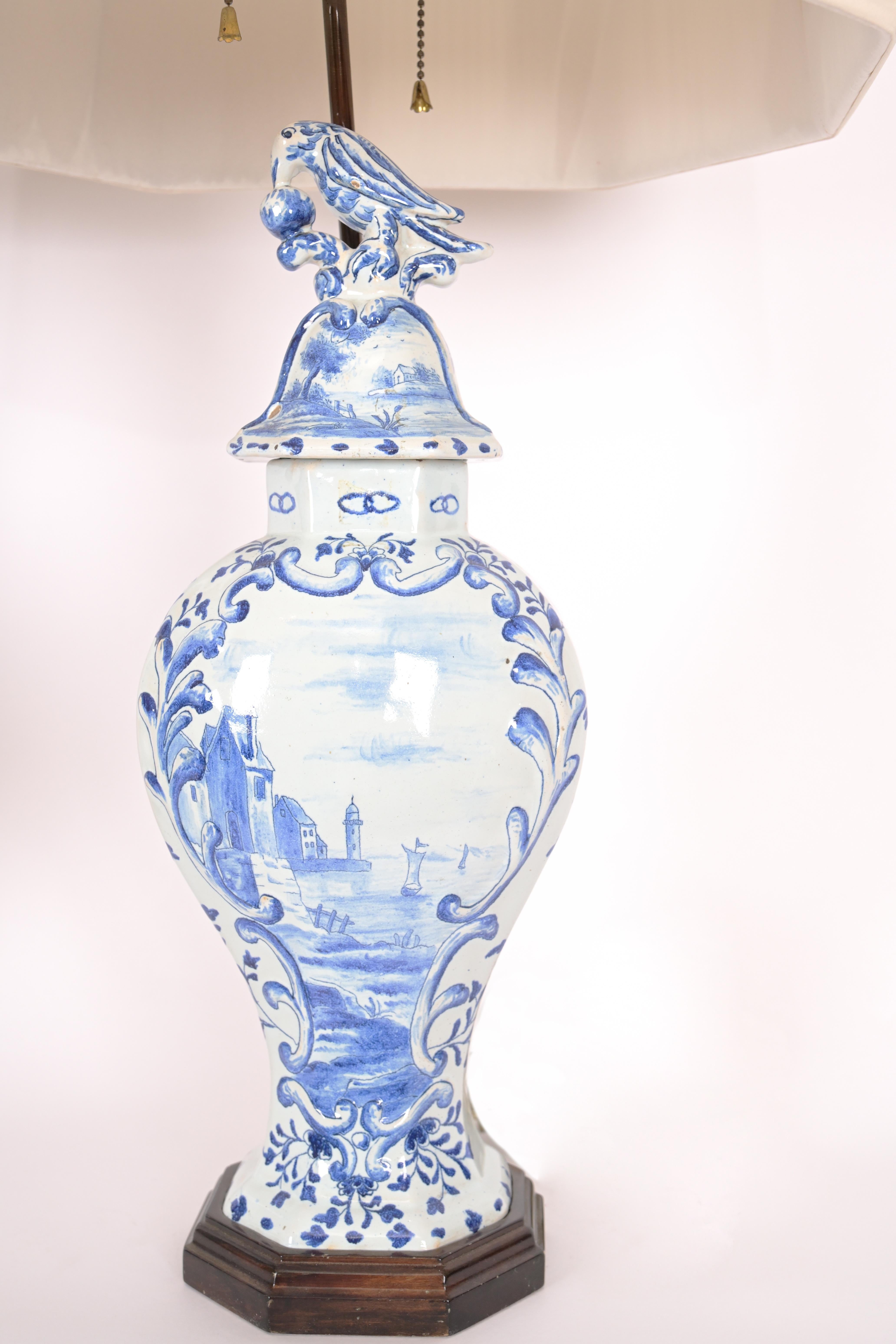 A pair of 19-century delft vases with traditional decoration with blue on white with mounted black painted bases and birds perched on the lids. Lamps mounted with special with brass wiring conforming to the silhouette feature. Now electrified with