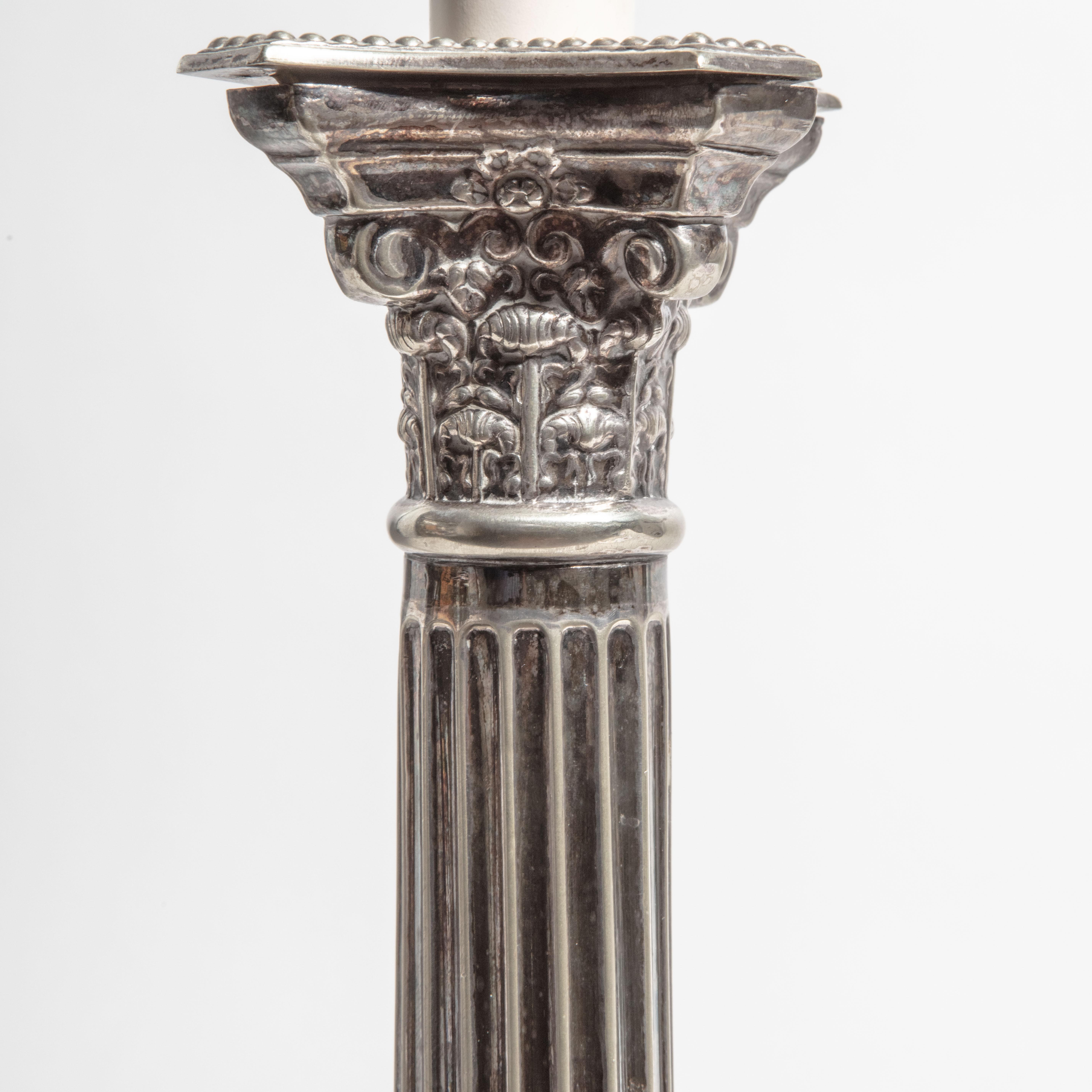 Pair of silver plate Corinthian column candlesticks, with Corinthian capitals above fluted stems and raised on stepped square bases. Now converted to lamps.

Measures: Height to top of column 32.5cm 13in
Height to top of shade 69cm 27in.