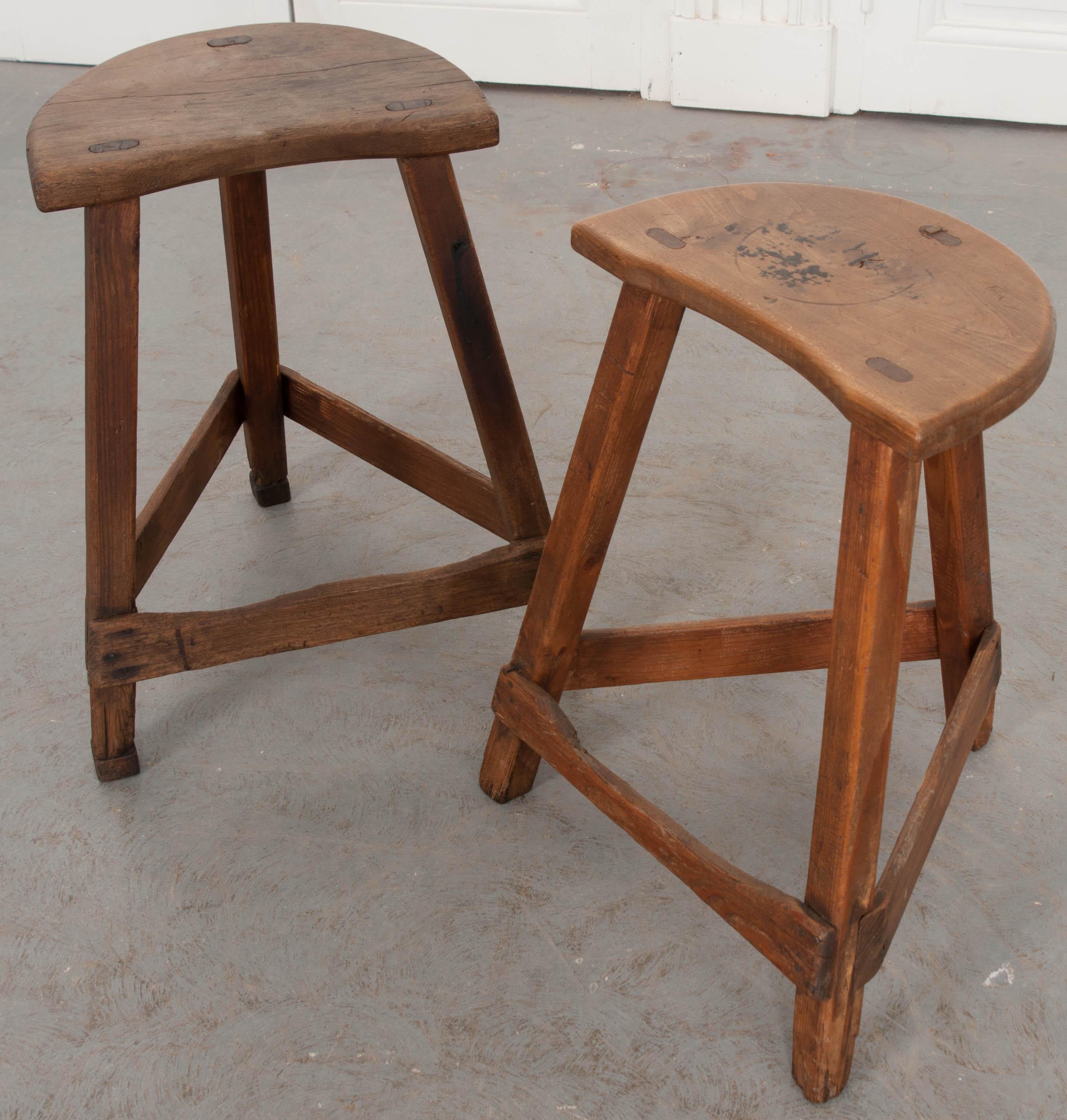 A charming pair of 19th century English low farm stools, made with elm seats and pine bases. Each stool has semicircular shaped elm seats that are supported by tripodal bases. The bases are made of pine, with their front braces markedly worn from