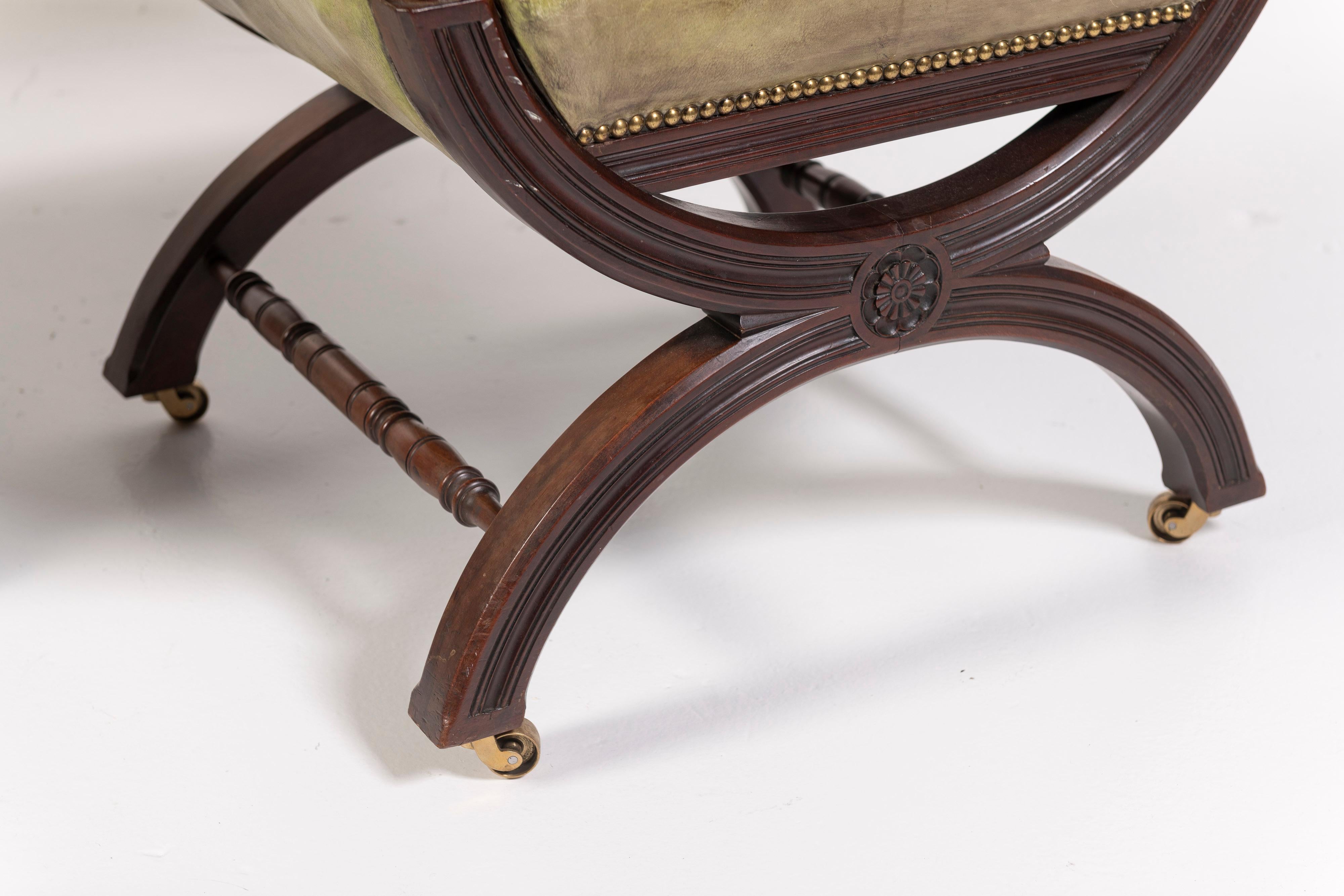 This fantastic pair of 19th Century English arm chairs, with a variation of the x-frame design, are made of mahogany and upholstered in an olive green, patinated leather. The carved legs, hand-nailed decorative studding in brass and the distinctive