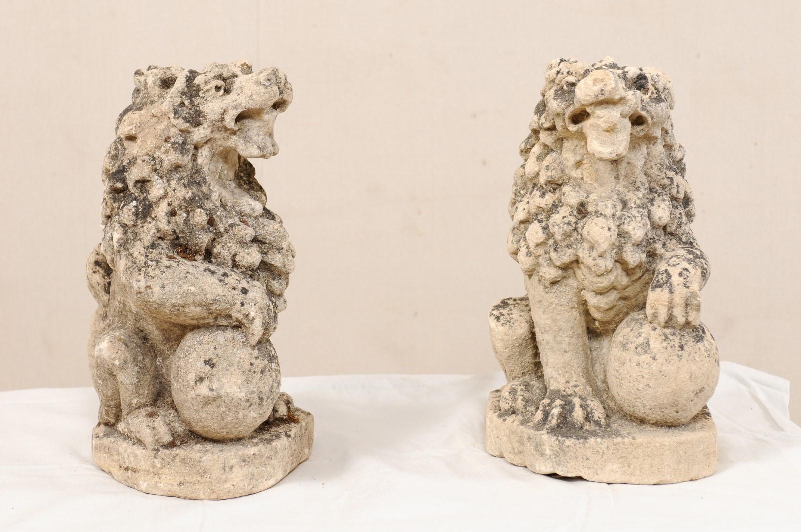A pair of 19th century English lions from carved limestone. This antique pair of stone lions from England are each shown in seated position with a single front leg or paw raised atop a ball. This pair of lions have gorgeous, full manes of hair and