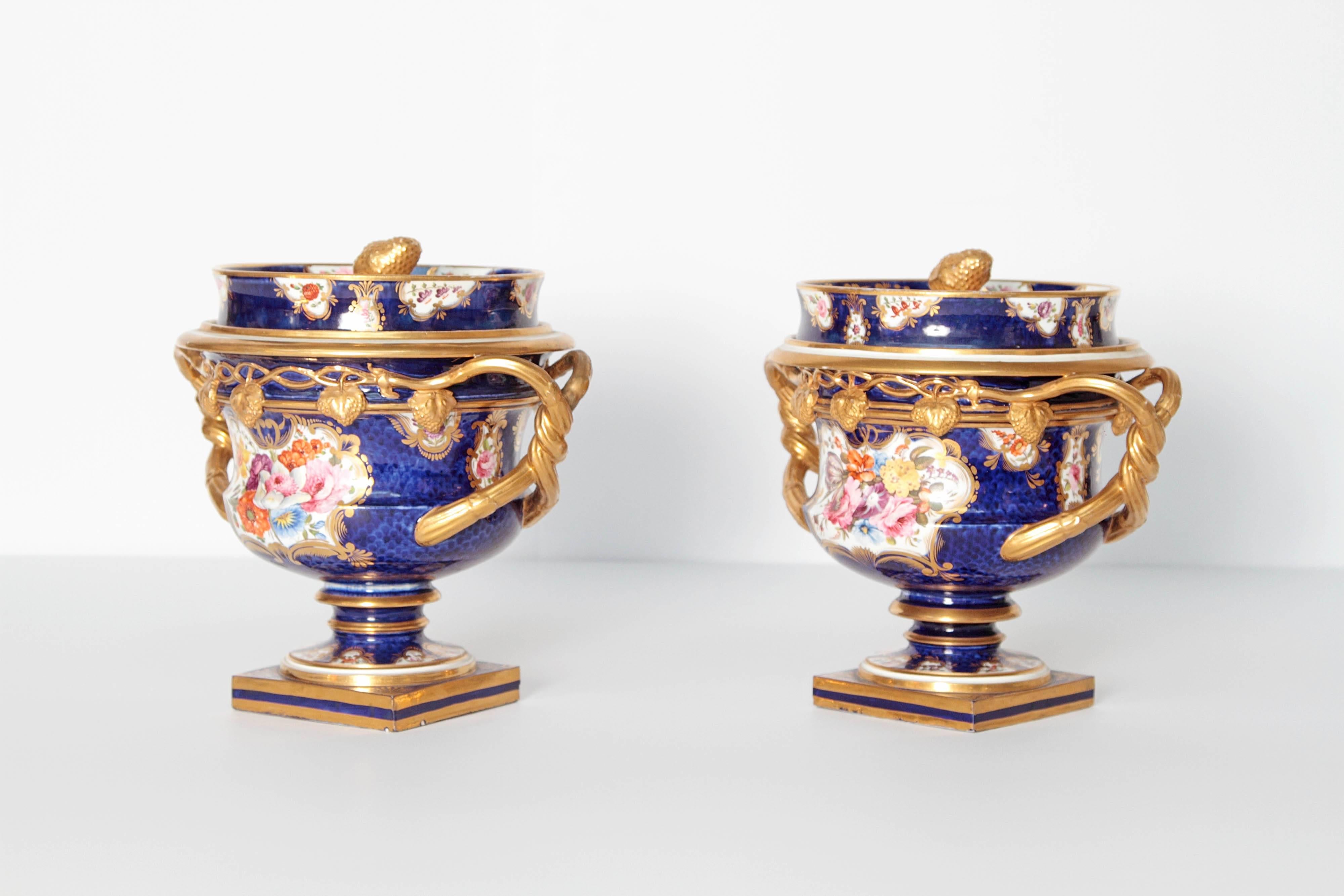 A pair of English porcelain fruit coolers with covers. Square plinth base and pedestal supporting bowls with twisted gilt handles, one of which appears to have been repaired / professionally restored. The covers with waisted gallery and strawberry