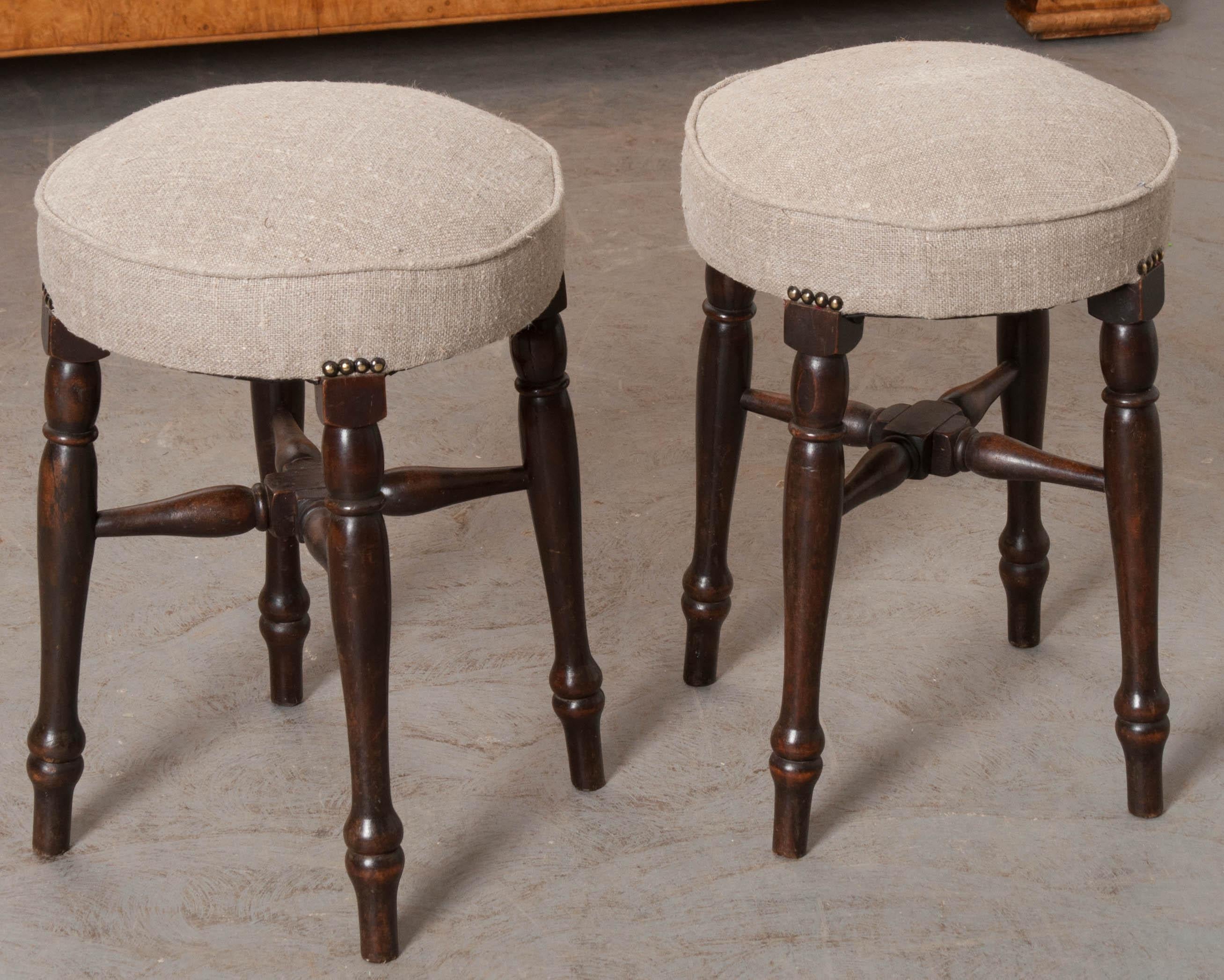 A fun pair of upholstered English stools, made in England, circa 1890. They both have baluster-form turned mahogany legs that are braced by turned mahogany stretchers. The pair have been recently upholstered in a natural slubby linen with single