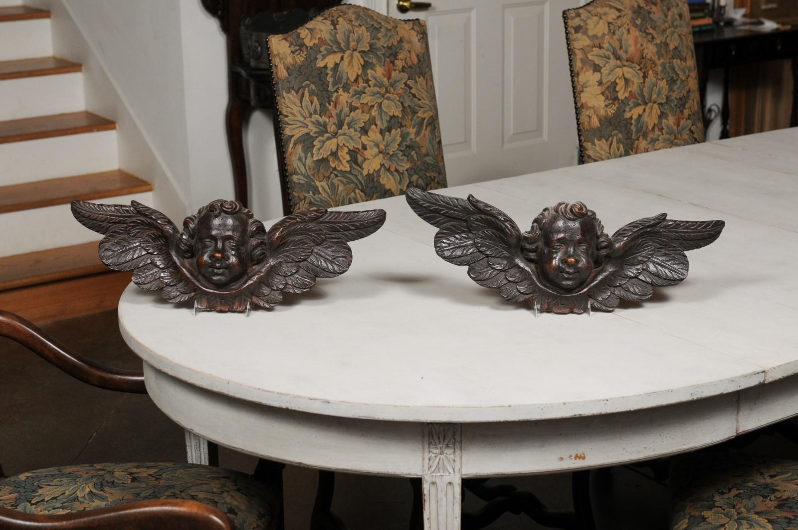 A pair of French Baroque style carved wooden angels from the 19th century with dark brown patina. Created in France during the 19th century, each of this pair of wall sculptures depicts a chubby angel head with curly hair, flanked with extended