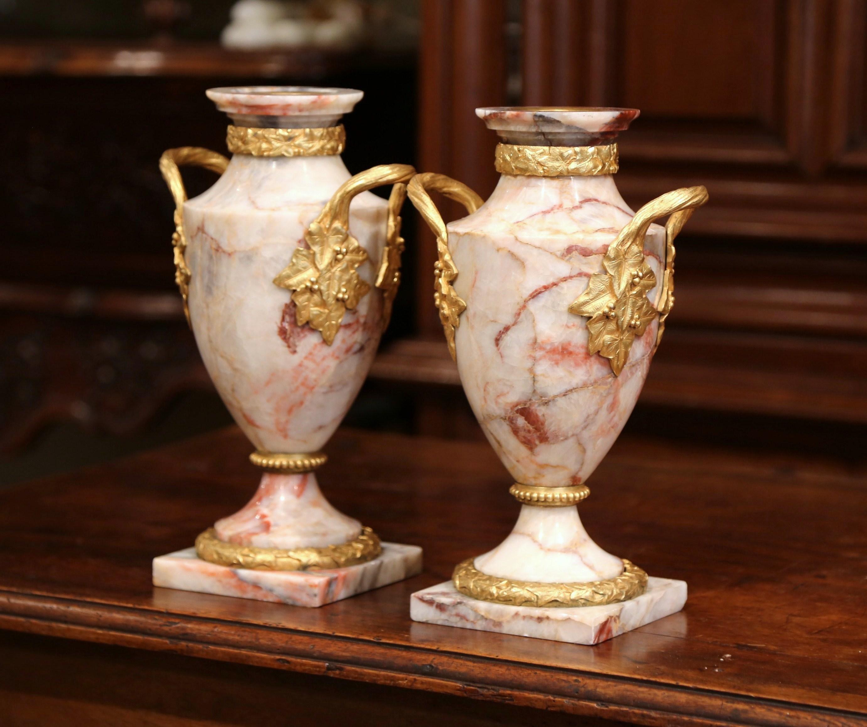 Decorate a mantel with this elegant pair of antique beige marble and bronze doré cassolettes urns. Created in France, circa 1870, the round carved marble urns with bronze handles stand on square bases, each vase is further decorated with ornate gilt