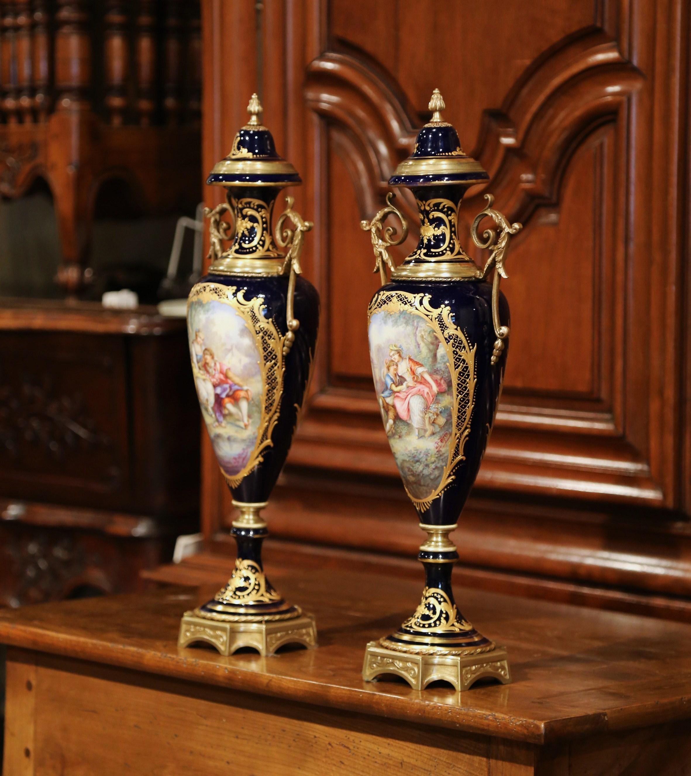 Decorate a mantel or a console with this elegant pair of porcelain and bronze urns with lids. Created in Sevres, France, circa 1880, each impressive lidded vase sits on a square bronze base and features hand painted romantic scenes over a signature