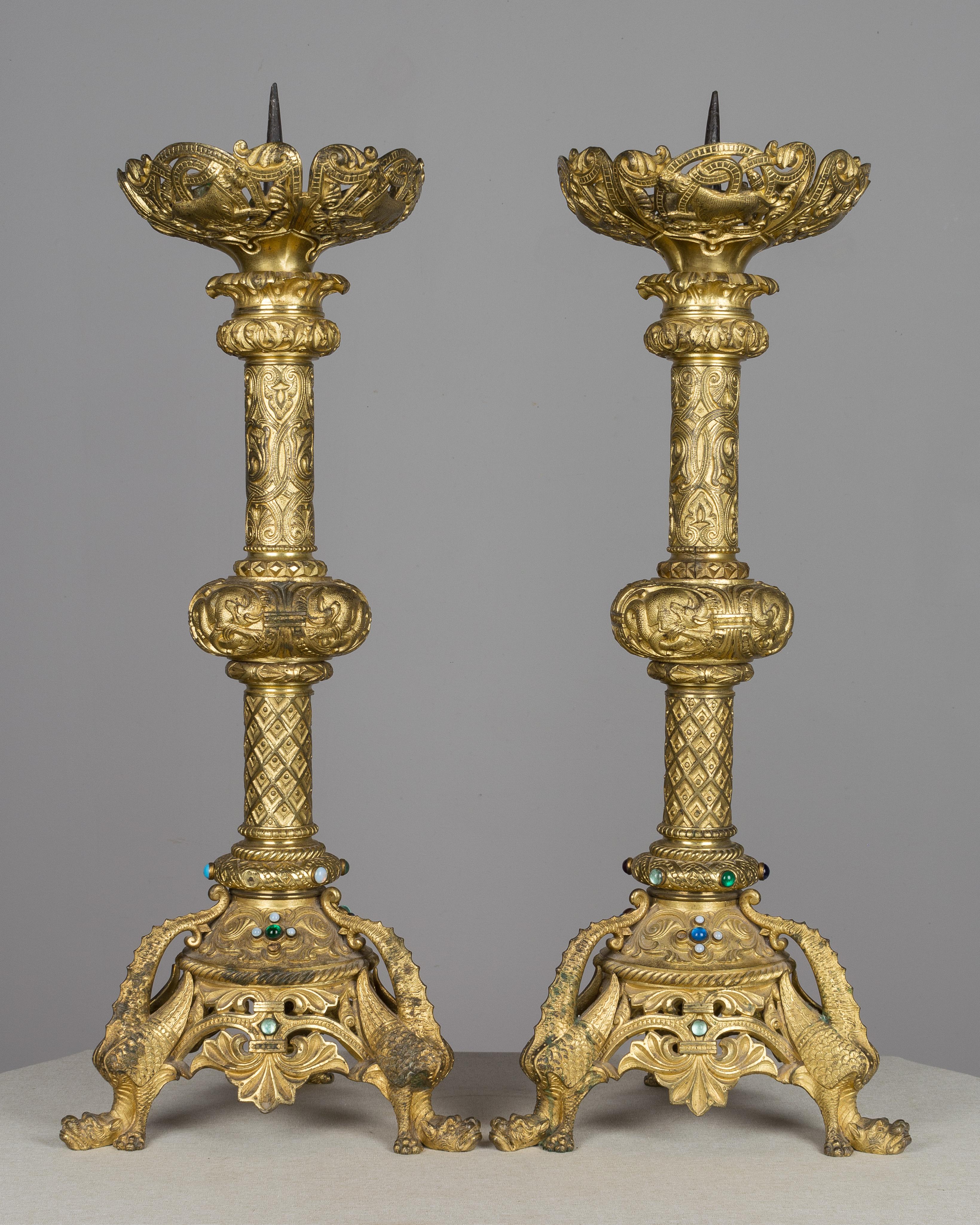 A pair of large ornate French bronze doré candlesticks. Heavy cast bronze with pierced decoration and set with colorful cabochon jewels. Triangle base with fantastic winged dragons. Large prickets for pillar candles and deep cups to catch melting