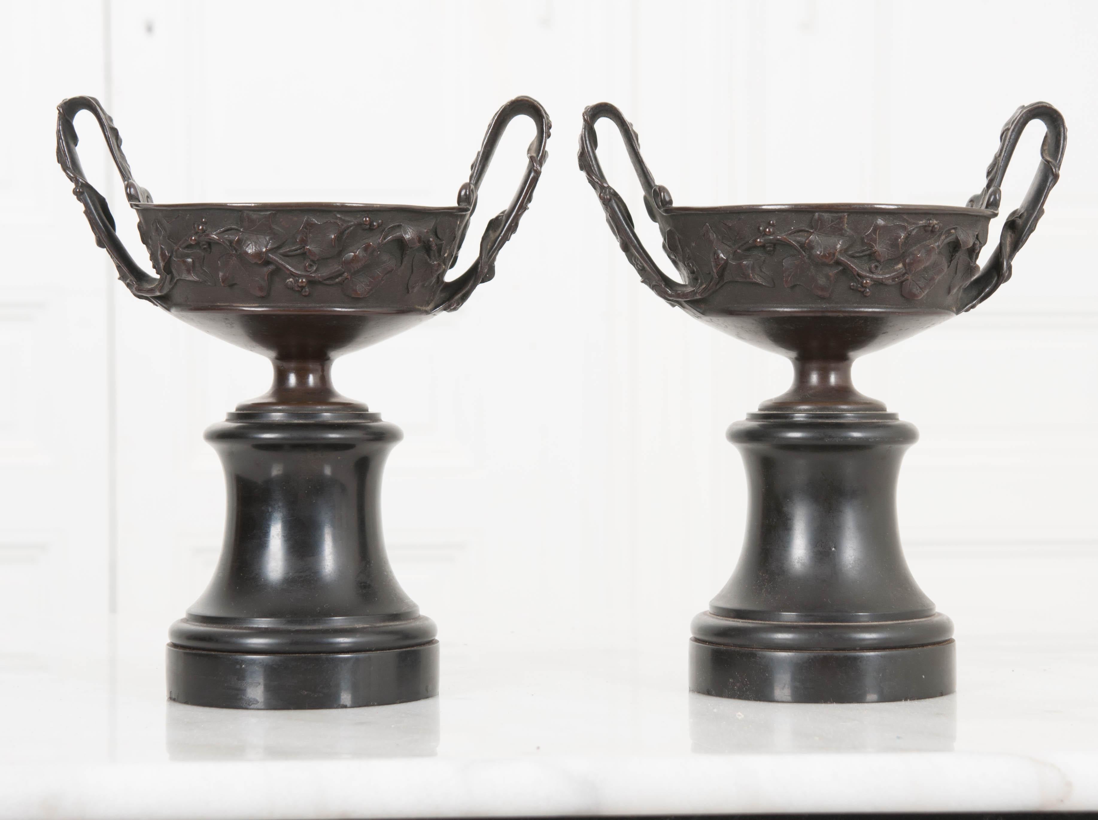 A lovely pair of bronze tazza-shaped urns from 19th century, France. The urns are made of cast bronze, and rest atop round, black marble bases. The bronze urns have been cast with a beautiful grape vine motif that extends around the bowl and up the