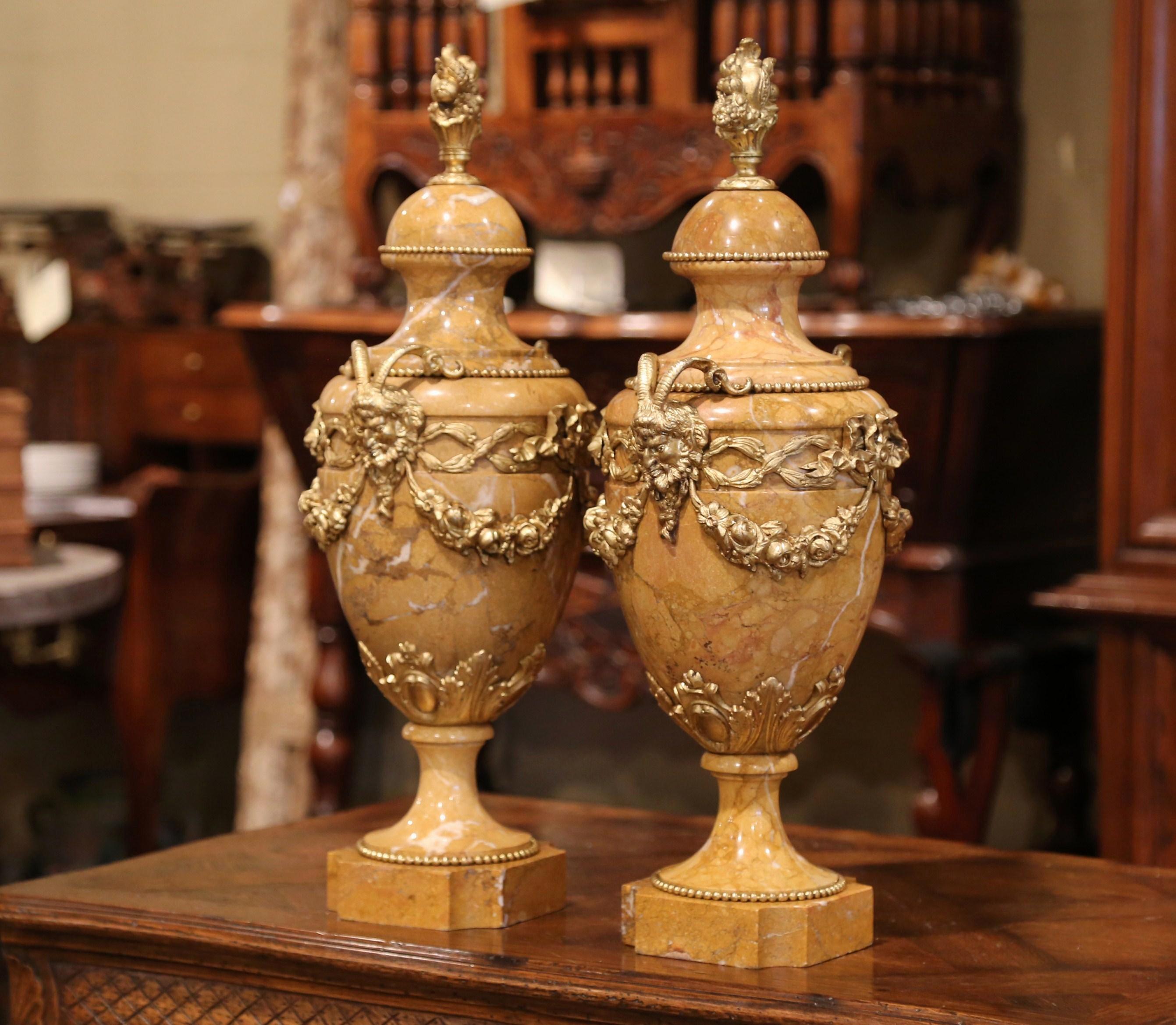 Crafted in France, circa 1870, this cassolette pair is hand carved from a beige marble stone and outfitted in bronze. The urns feature a decorative bronze finial at the top decorated with a fruit basket. The two side handles depict Bacchus (or