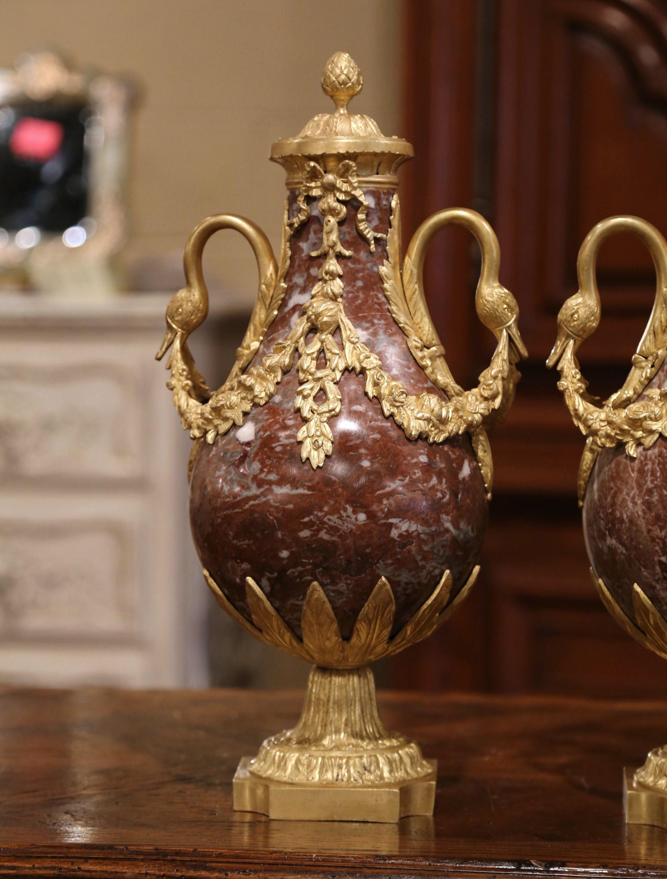 Crafted in France, circa 1870, these antique urns are made of marble and stand on square bronze bases decorated with leaf motifs. Each marble vessel features a decorative cone-shaped bronze finial at the pediment, a pair of bronze side handles with
