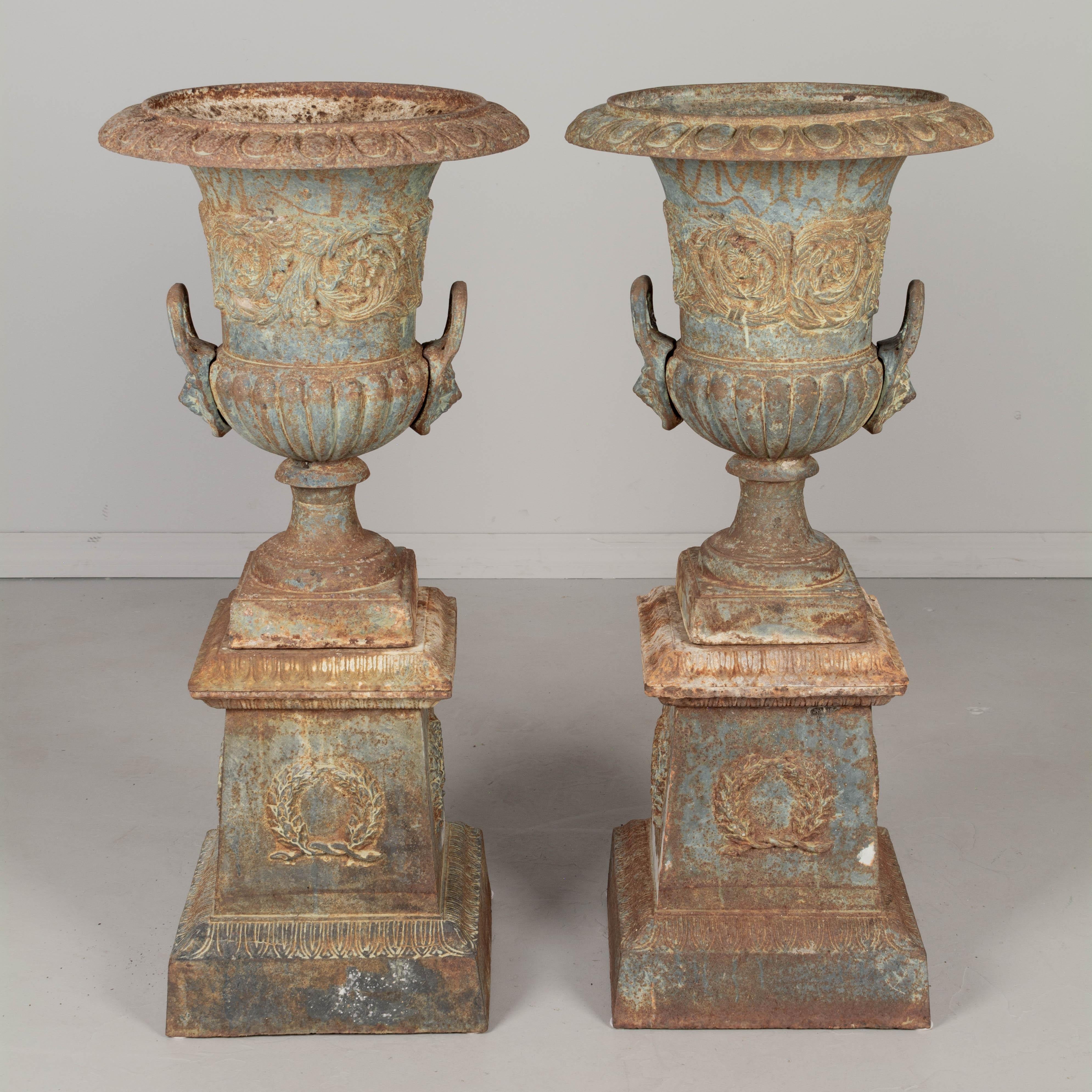 A pair of 19th century French cast iron garden urn planters. In three parts, the tall urn rests on a pedestal base which has a removable rim. Fine cast details with large lion head handles and swirling foliate relief across the face of the urns and