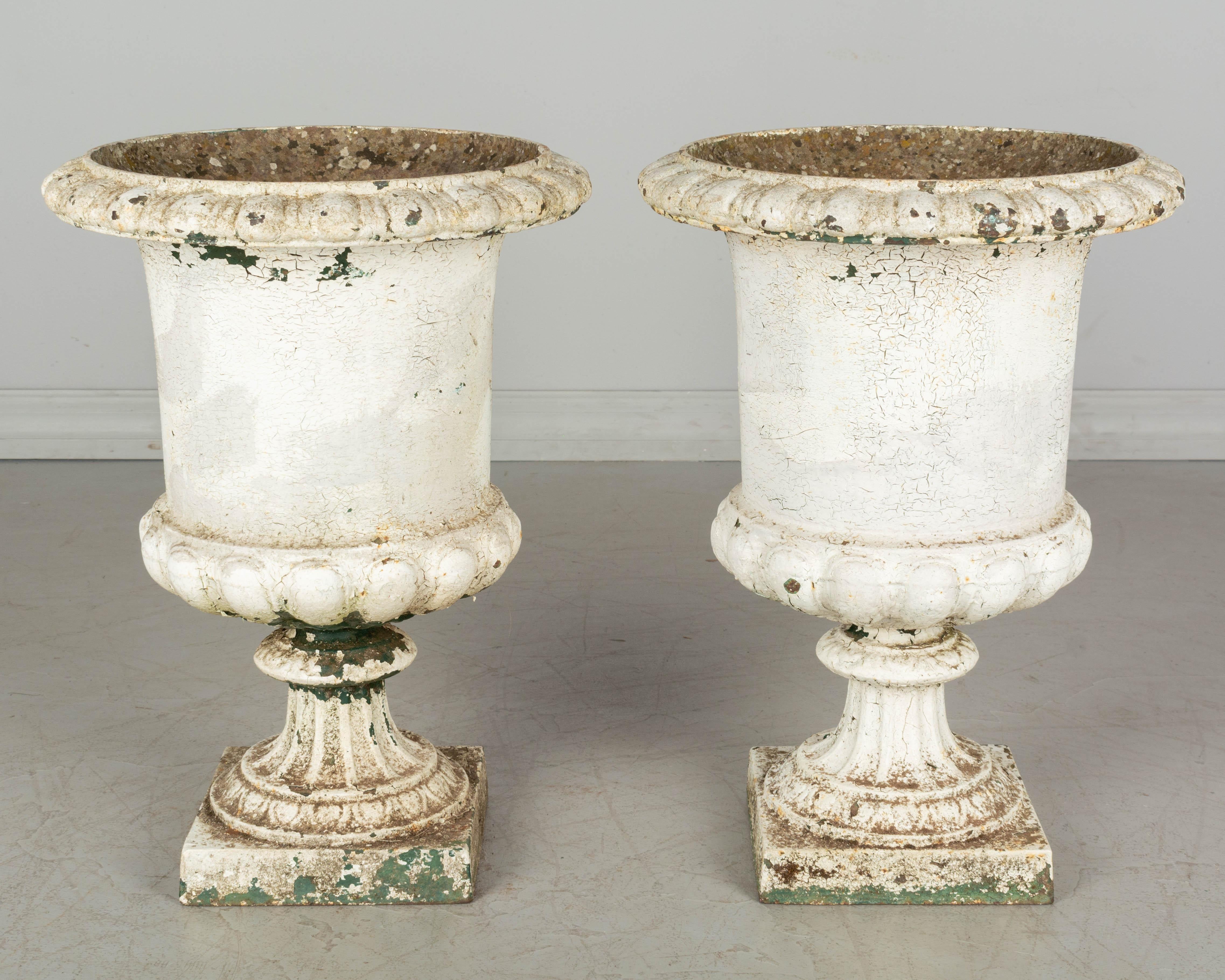 A pair of early 19th century French cast iron garden urn planters, with tall proportions. Original weathered white painted patina, cracking and peeling away is places exposing a layer of green paint beneath. Weight: 53 lbs. each. Measures: 19.75