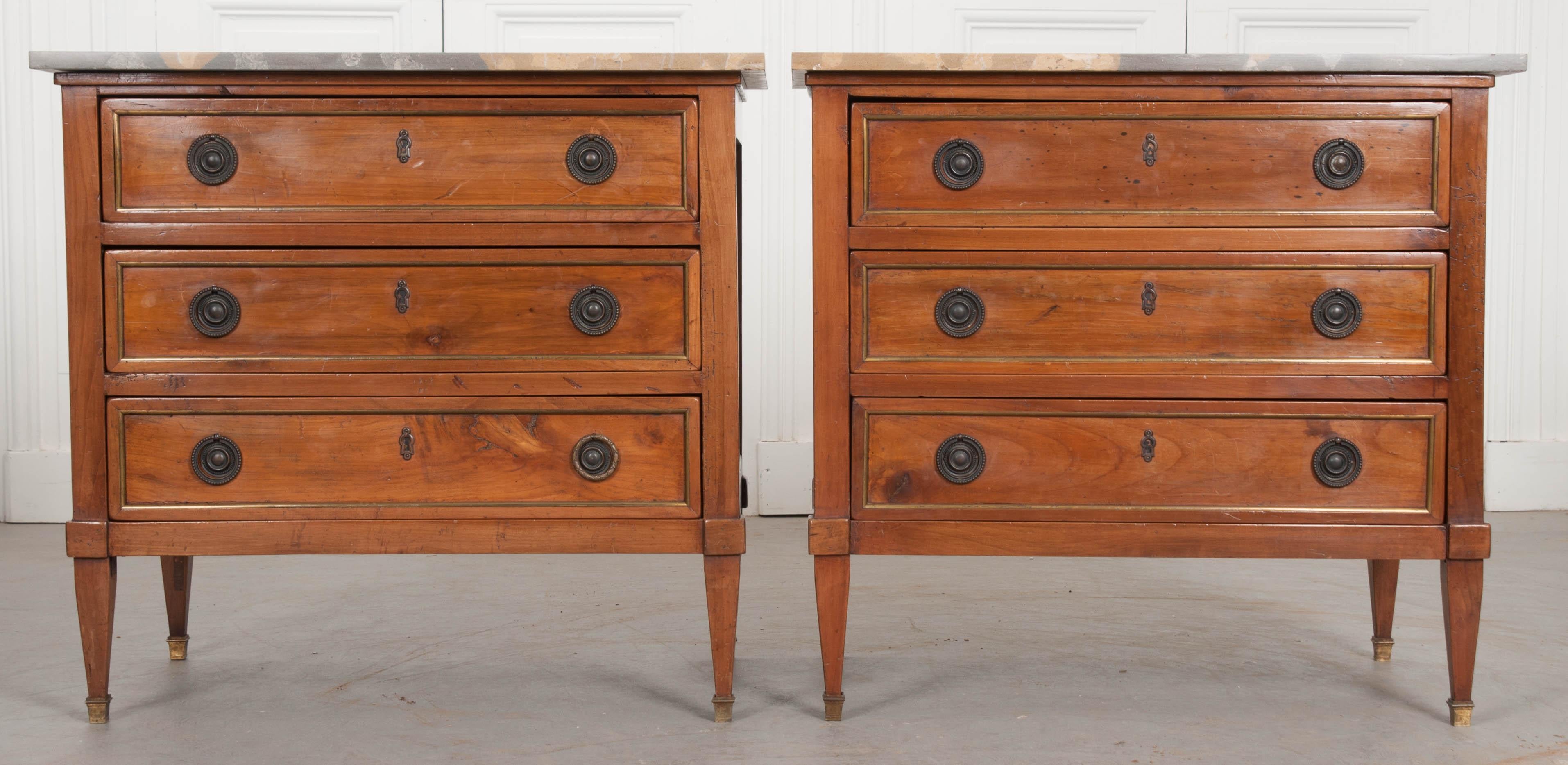 This stylish pair of Directoire-style commodes are from France, circa 1860, and feature beautifully unique gold-and-grey marble tops above a bank of three paneled drawers with round bronze ring pulls and escutcheons, and raised on square tapering