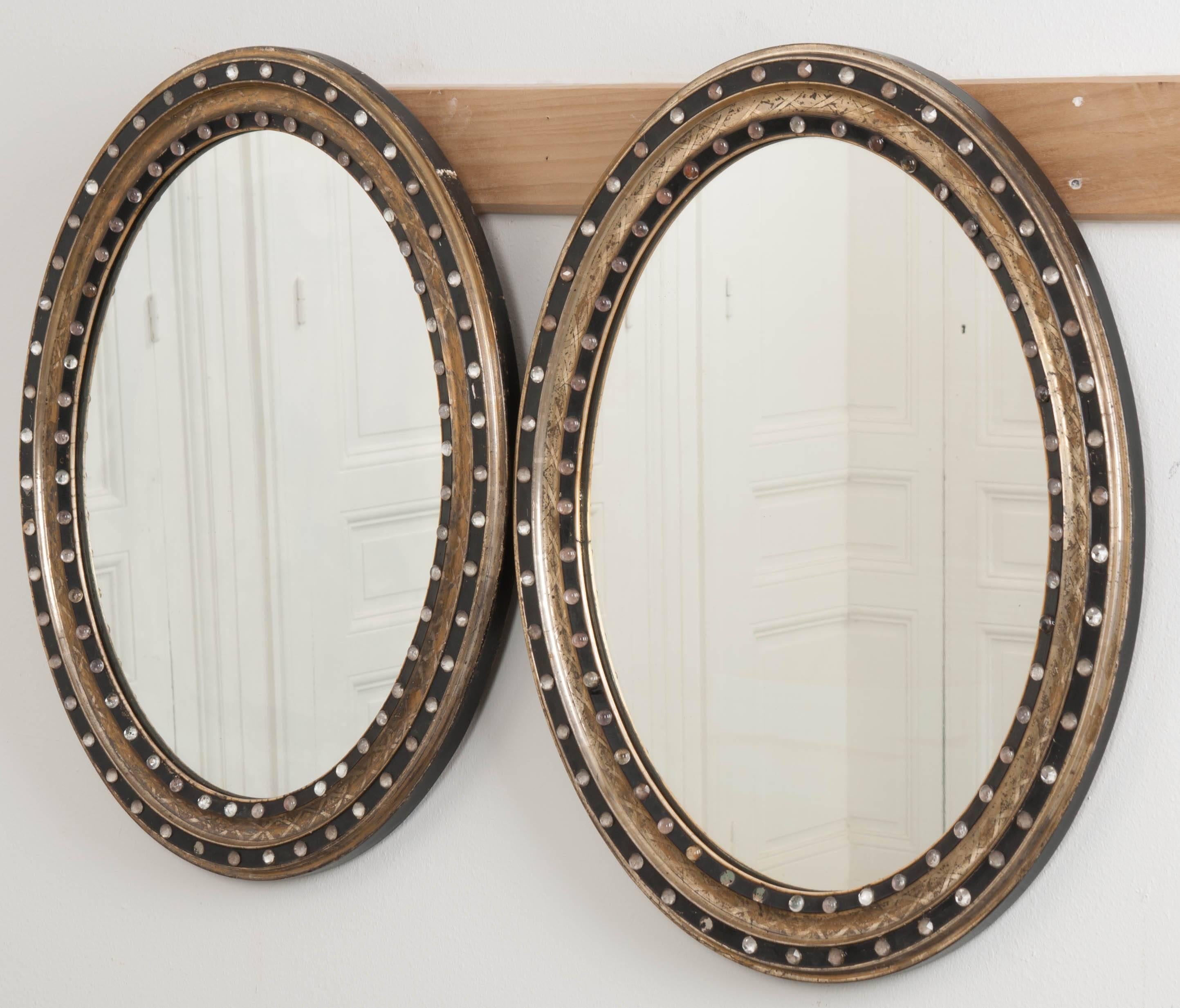 A wonderfully sized French oval mirror that is uniquely styled. Combinations of silver and gold metallic gilt sharply contrast the ebonized frame which has been decorated with round and faceted crystals. Crosshatched etching, along with the inlaid