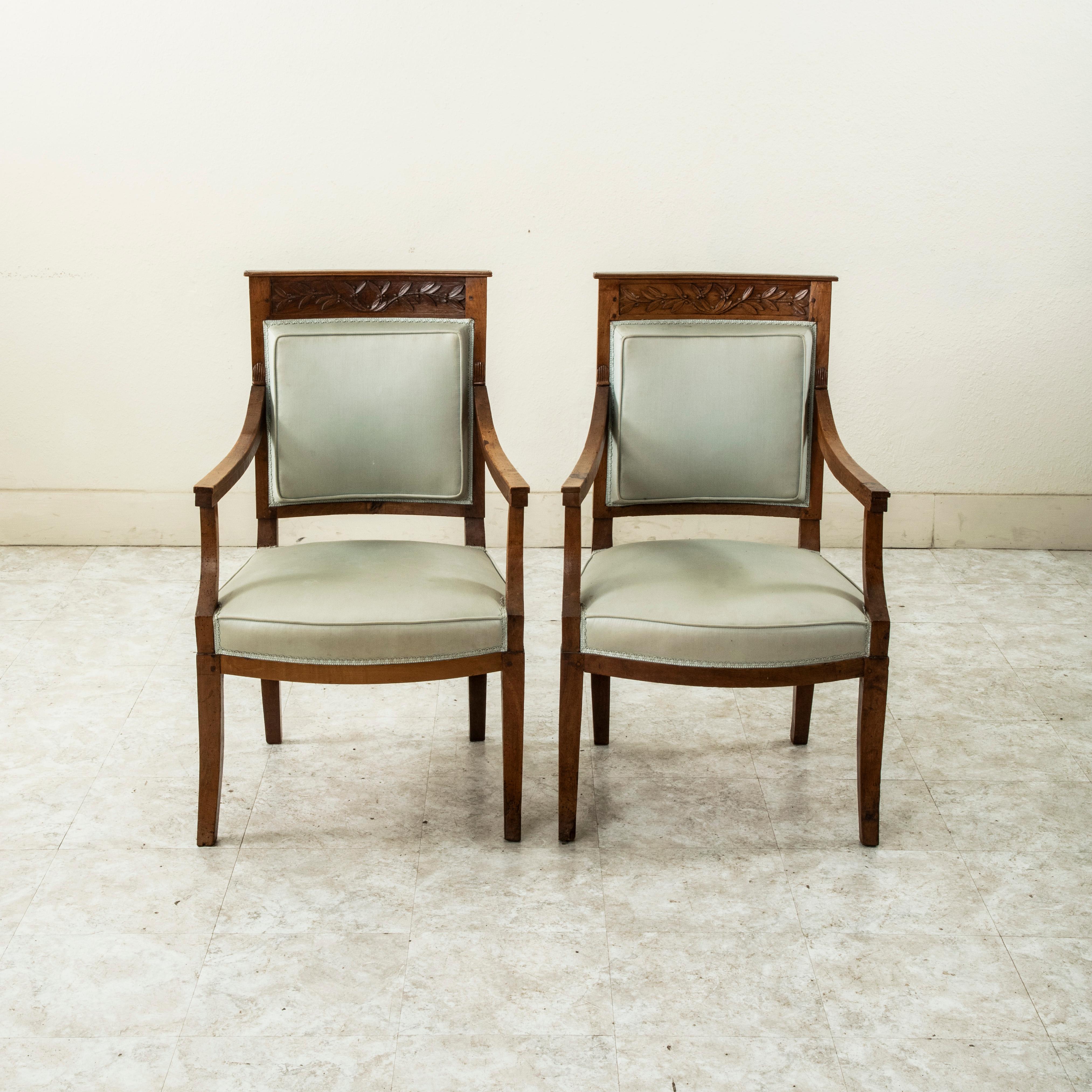 Cette paire de fauteuils en noyer de style Empire français du début du XIXe siècle présente des détails sculptés à la main de lauriers sur le dossier de l'assise et de coquilles au sommet des accoudoirs. Les dossiers sont incurvés pour plus de