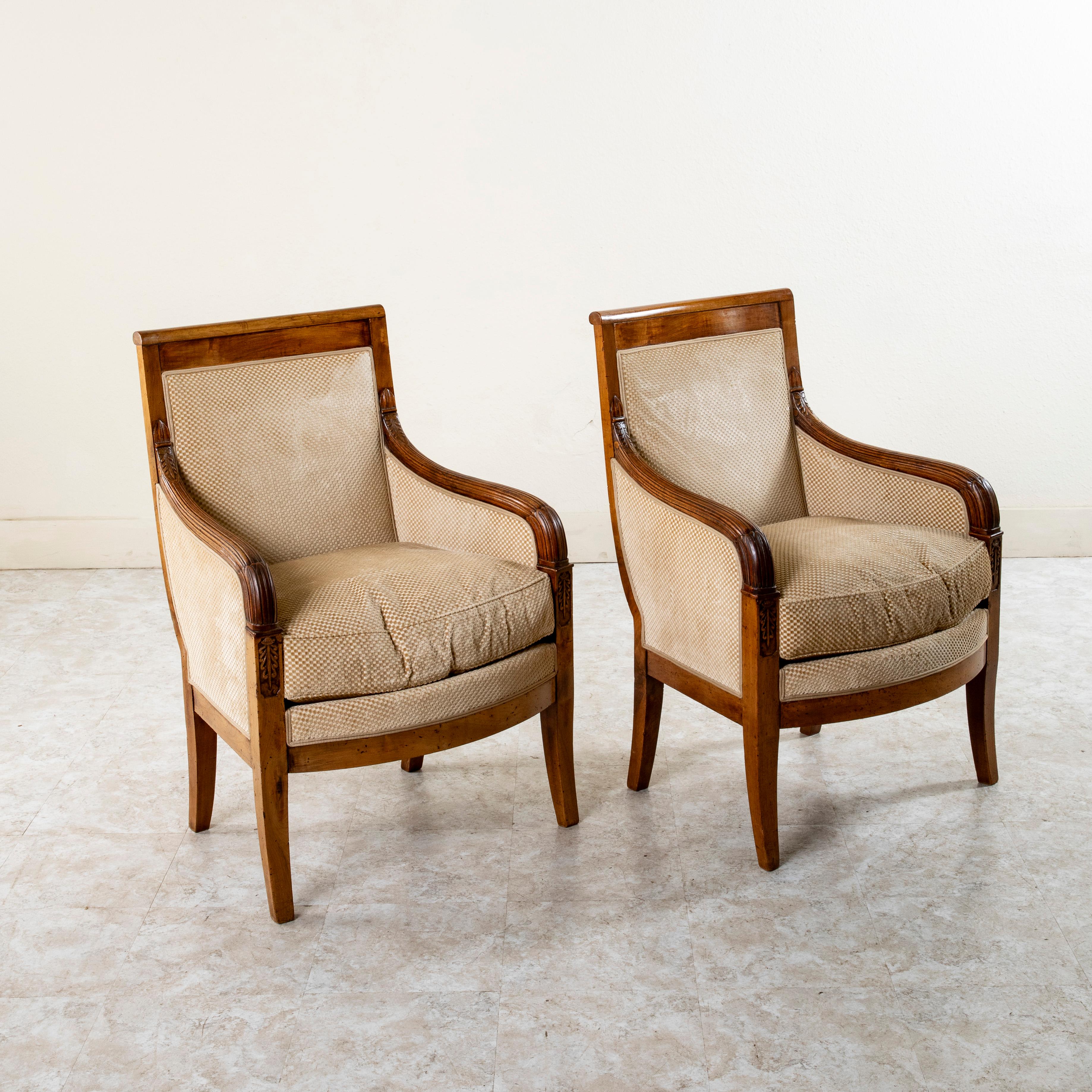 From a home in Versailles, France, this pair of early nineteenth century French Empire period bergeres or armchairs is constructed of solid walnut with a rich patina that only comes with age. These chairs feature beautifully hand carved armrests