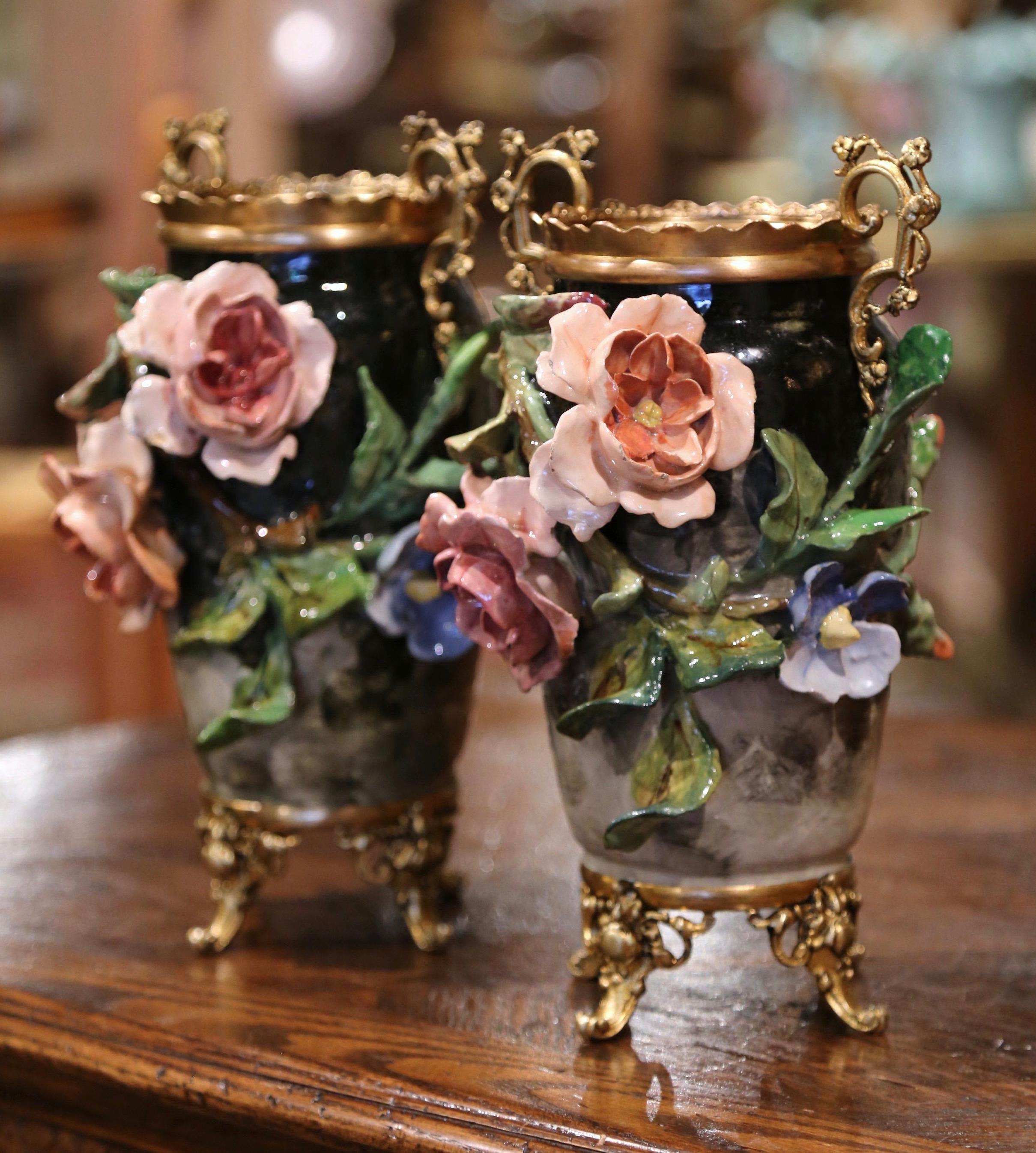 Decorate a kitchen counter or display a shelf with this elegant pair of antique Majolica vases. Crafted in Montigny sur Loing, France circa 1860, each ornate vase sits on a brass base with scrolled feet, and features colorful hand painted flowers