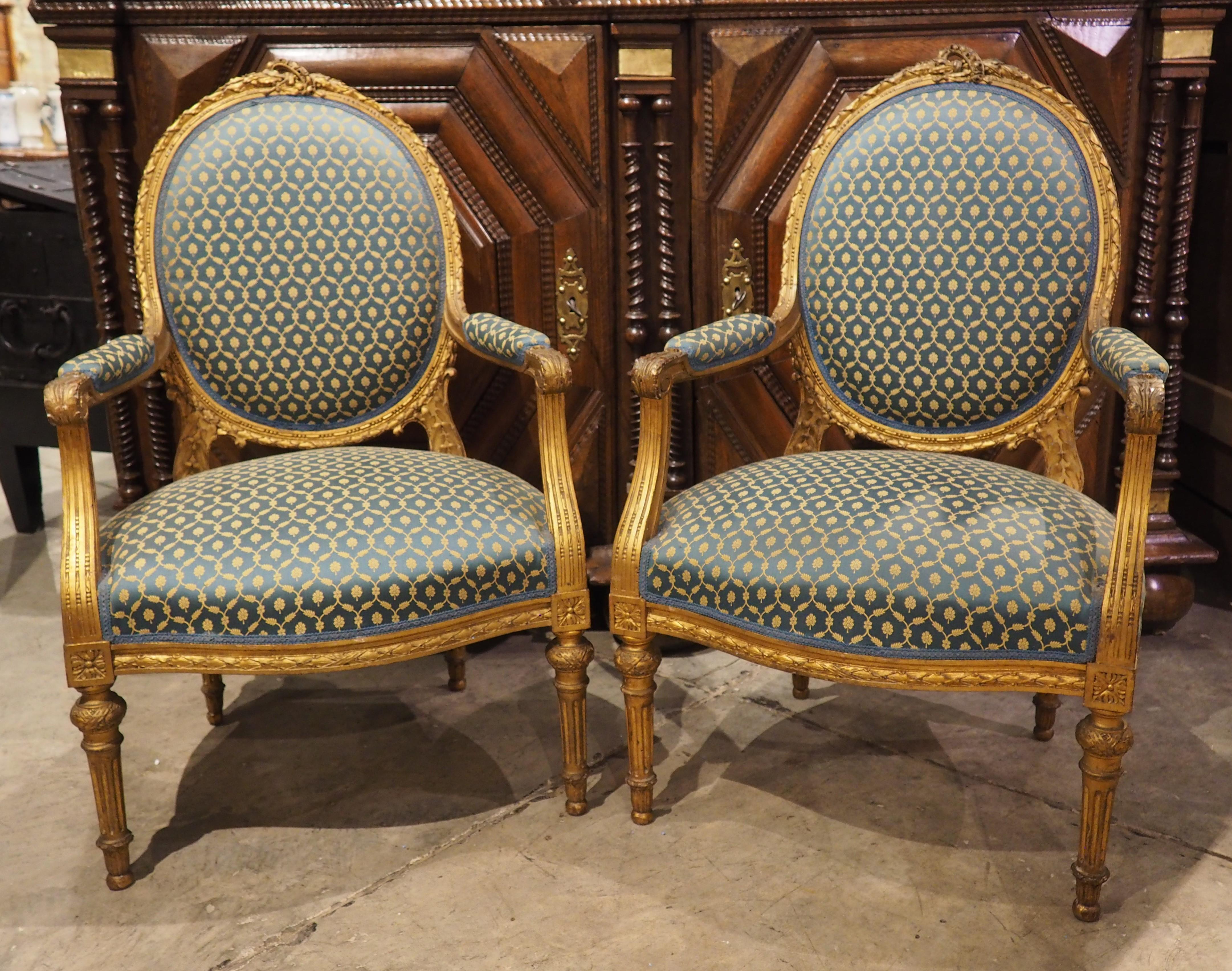 More information coming soon…

A pair of Louis XVI-style armchairs, hand-carved in France in the 1800s, each chair has a giltwood frame with several areas upholstered with a teal and guild fabric.  The oval medallion back is topped by laurel