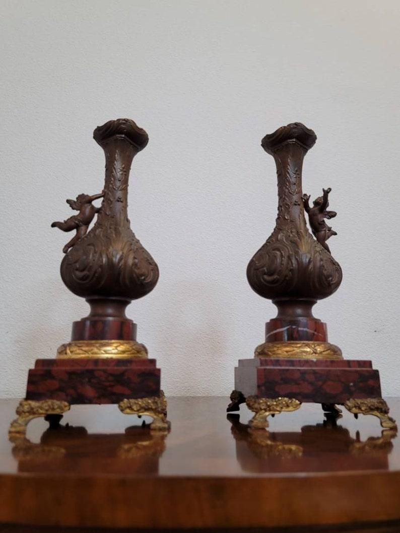 A pair of French patinated bronze, gilt metal and marble garnitures from the late 19th century.

Long bottle-neck vase form with foliate scroll design in relief, winged putto, Rouge Griotte marble base, on gilt cast metal bracket