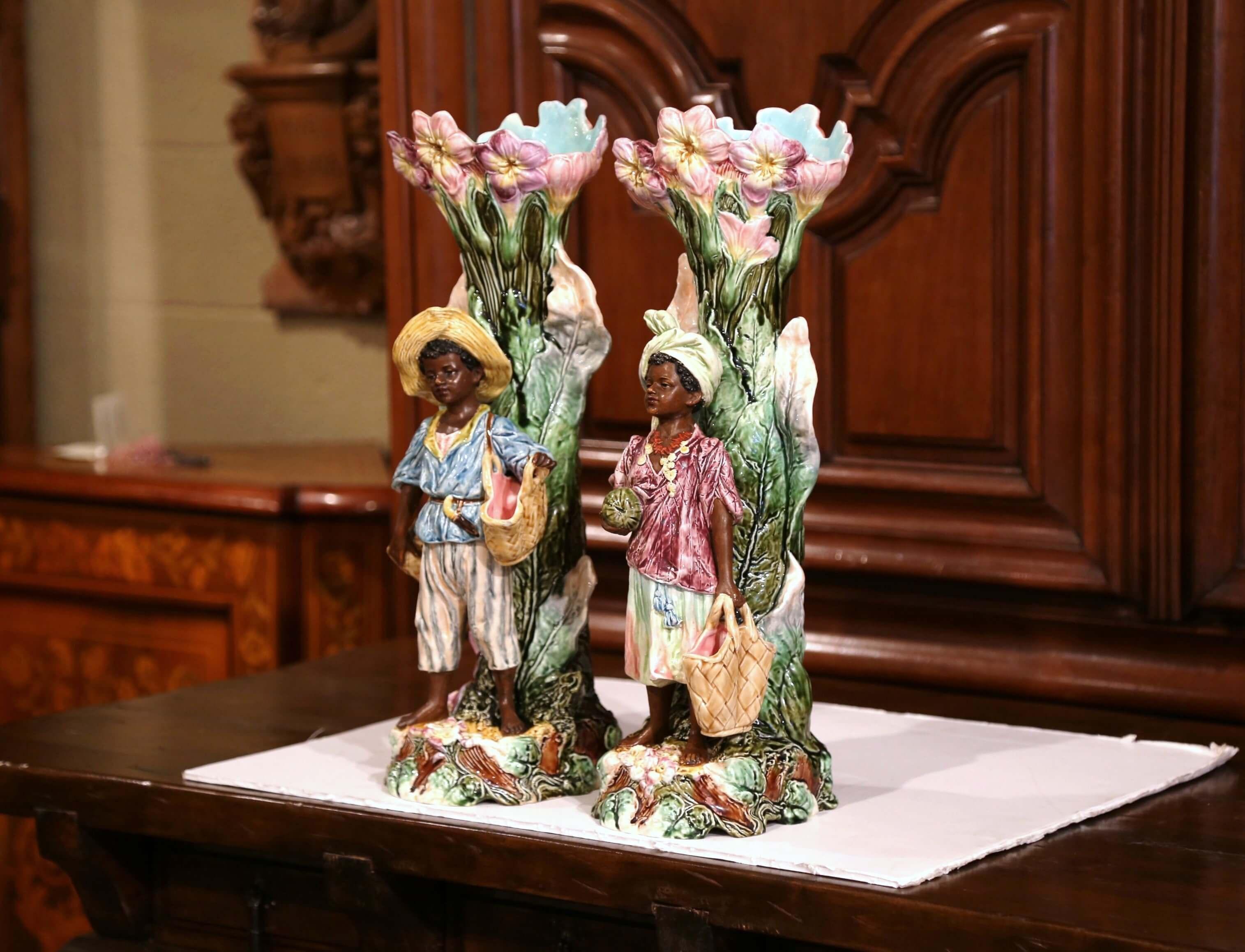 This beautiful, unique pair of antique Majolica vases was crafted in France, circa 1880. The tall and thin colorful ceramic planters feature two hand painted children in high relief returning from the market, embellished with floral decor. Each