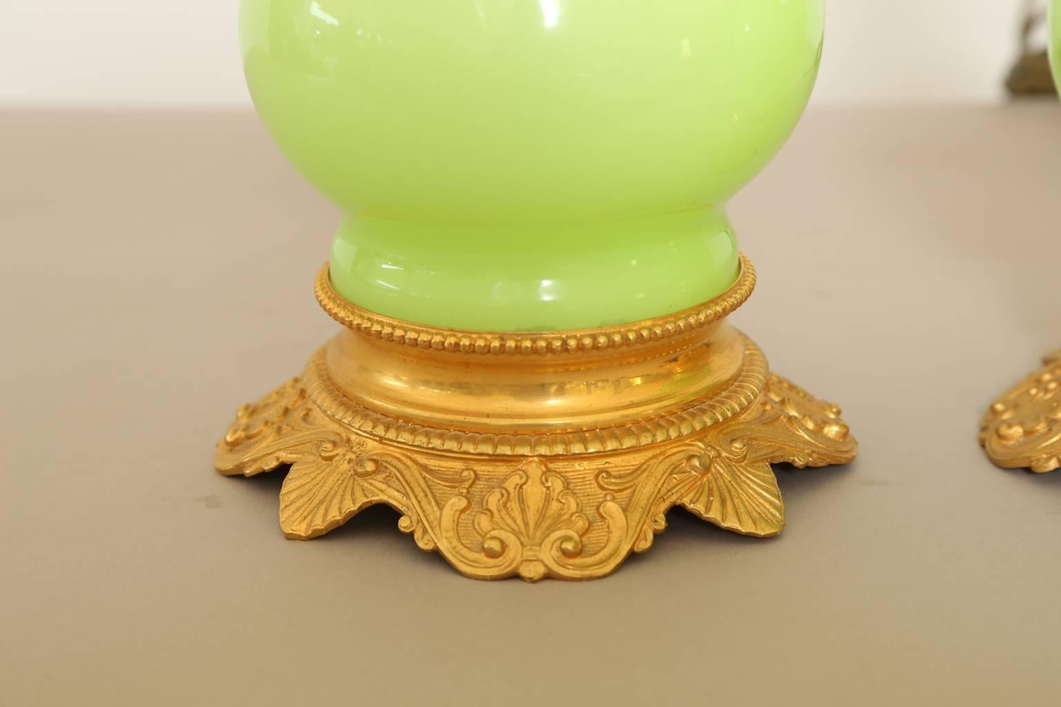 Pair of lamps, originally oil-burning, of lime-green opaline glass, each having a baluster form, with ormolu accents; lamped on serpentine base decorated with scrolling palmettes and scallop shells, its burner collar with detailed, pierced neck.