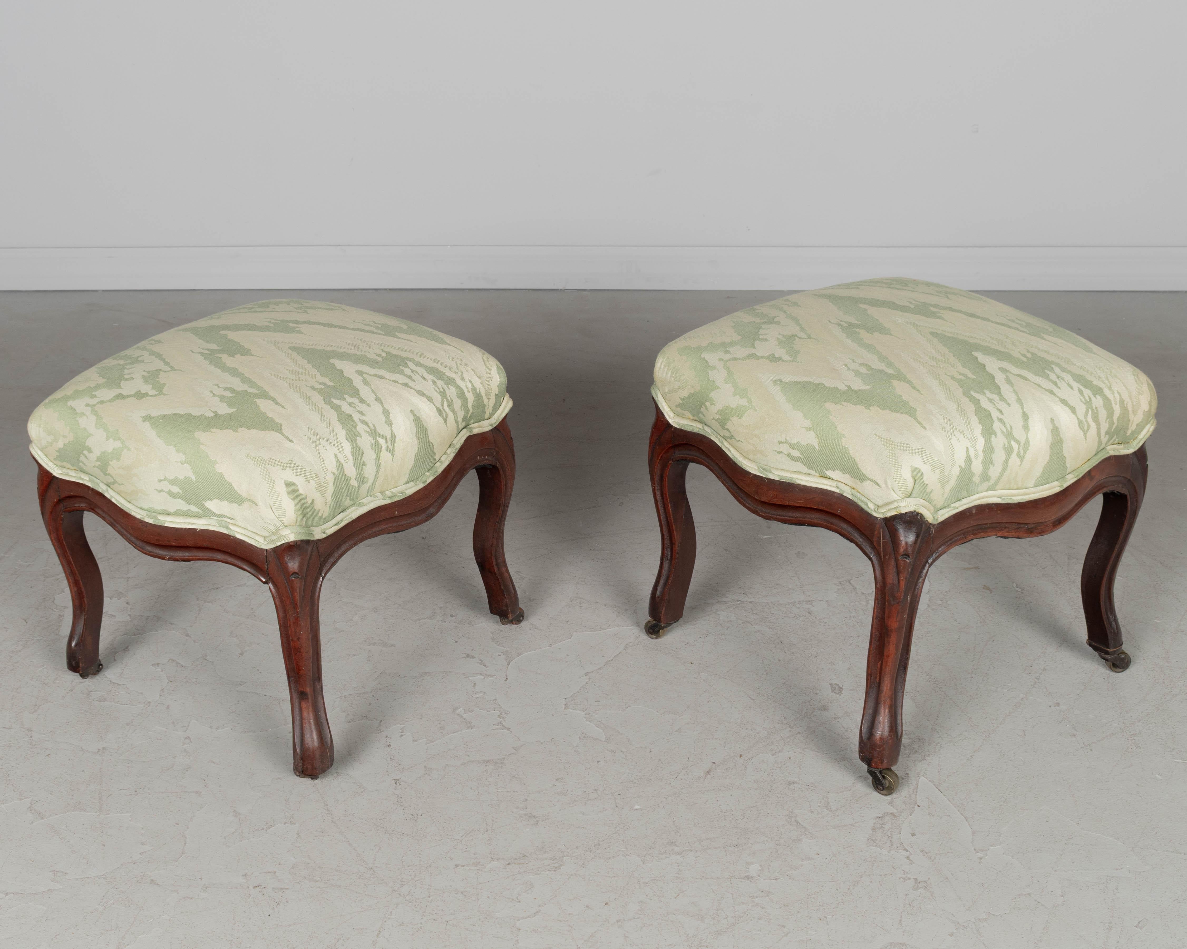 Pareja de taburetes franceses para los pies, estilo Louis Philippe, tapizados, con armazón de caoba tallada. Una con ruedas de madera y otra con ruedas de latón. Bien tapizado en tela verde pálido. En buen estado, con armazón robusto, algunas