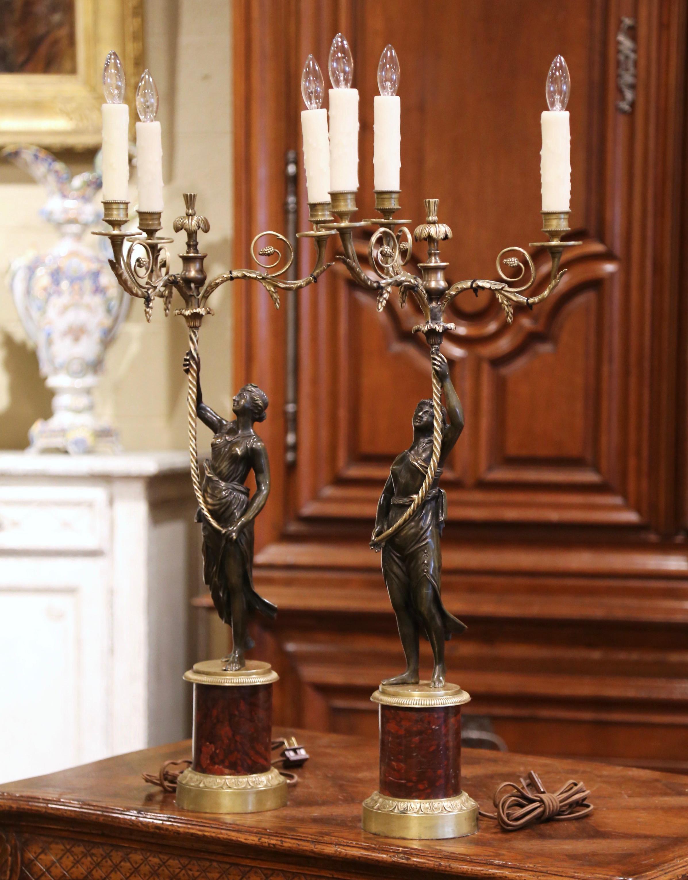 Crafted in France, circa 1880, each antique candelabra stands on a cylindrical marble plinth base, and features a bronze classical female figure holding a torch with three scrolled lights. Each tall table fixture is wired and dressed with real