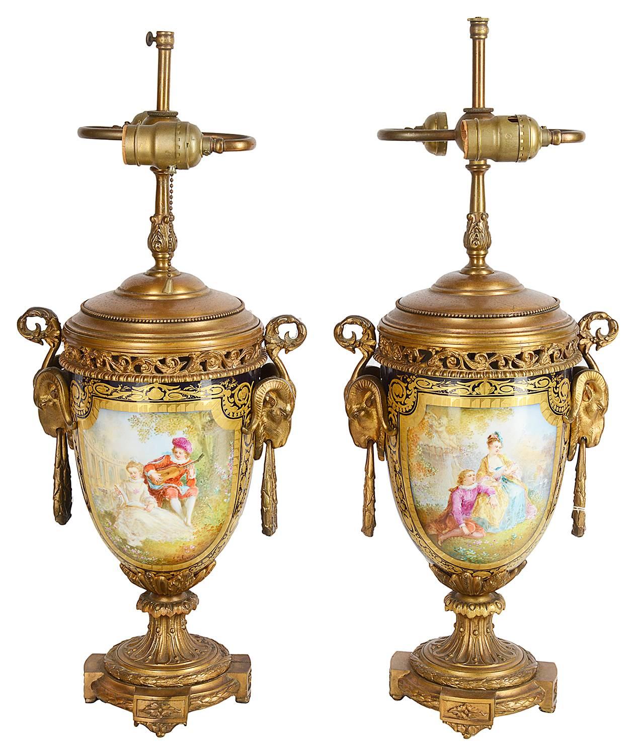 A good quality pair of French Sevres style, ormolu-mounted vases or lamps. Each with Rams head mounts, a cobalt blue ground with gilded decoration around painted panels depicting classical romantic scenes.