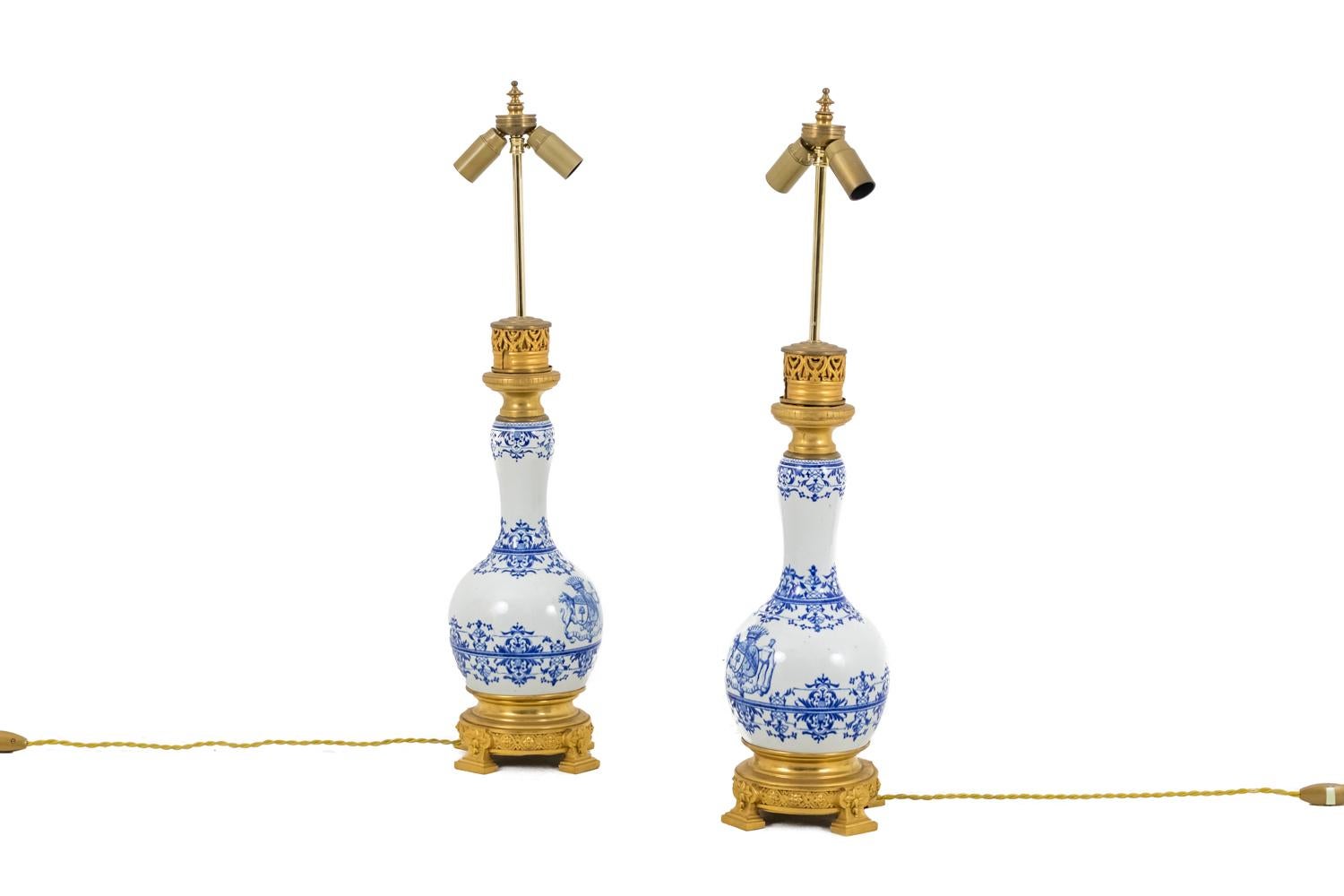 Pair of Gien earthenware lamps mounted in gilt bronze, standing on a circular base with rosettes interlacing.
Blue and white lambrequins and stylized vegetal decoration. Coat of arms with two shields underlined by the ribbon motto « Semper virens »