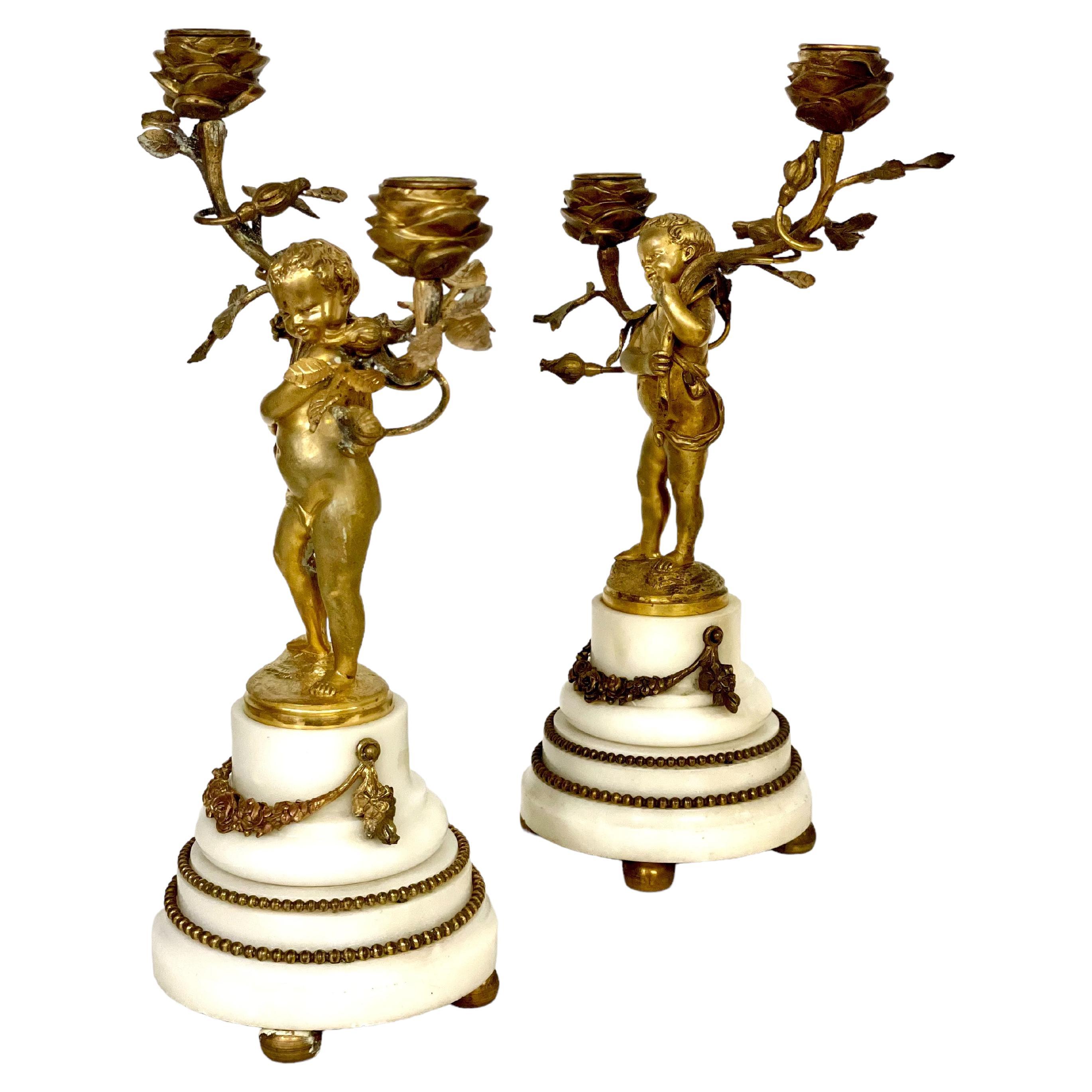 A fabulous pair of complementary 19th century gilt bronze and white marble two-branch candelabra, crafted in the Louis XVI style. Each of these ornate candle holders takes the form of a barely veiled golden putto holding aloft branches of billowing