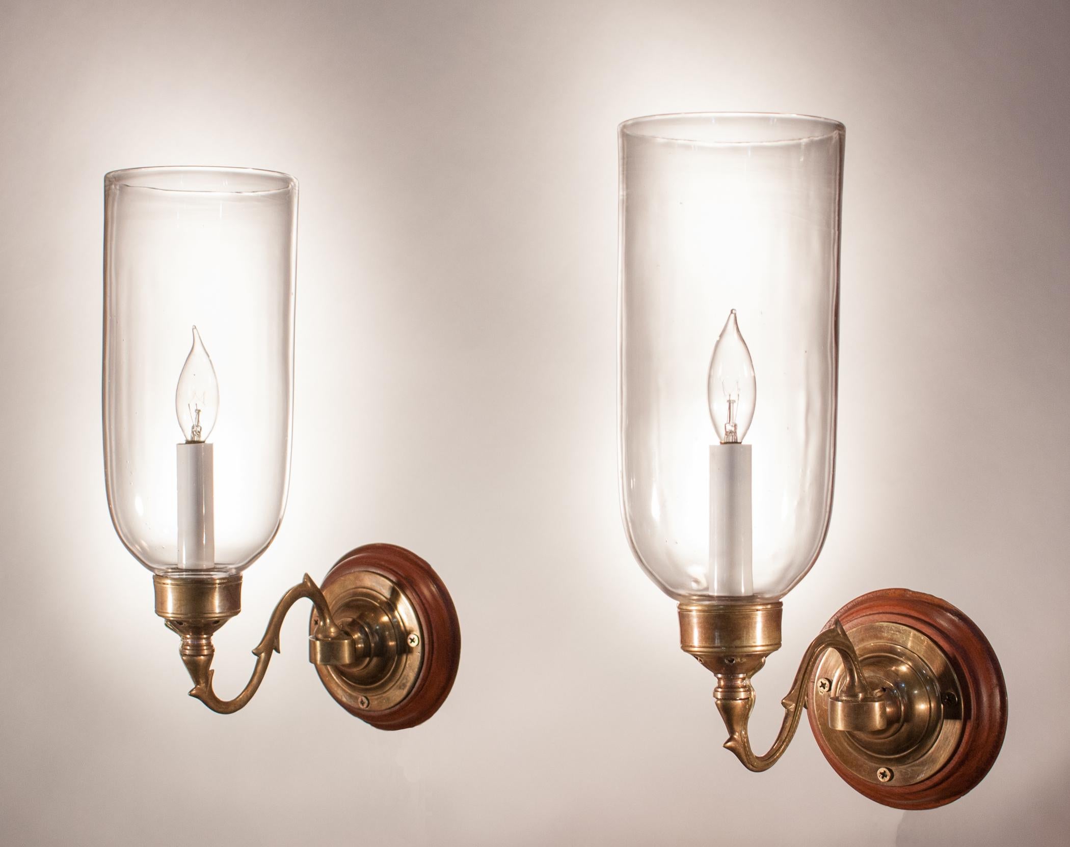 Classic pair of hand blown glass hurricane shades from England, circa 1890. The cylindrical shades have lovely form and their proportions are well-suited to these compact sconce arms. Custom-fabricated brass sconce arms and mahogany backplates (as