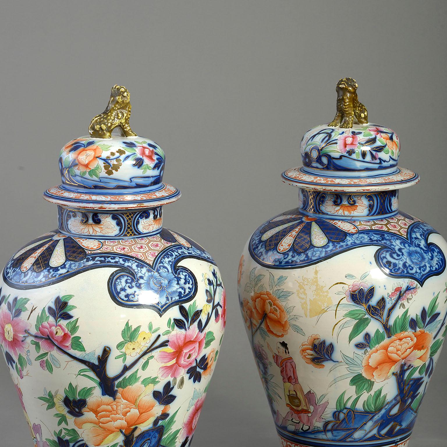Ein seltenes Paar Fayence-Vasen und -Deckel aus dem frühen 19. Jahrhundert, dekoriert im Imari-Stil, mit vergoldeten Löwenköpfen, die Körper reichlich mit Figuren, exotischen Vögeln, Blumen und Blättern verziert. Auf ebonisierten Holzständern