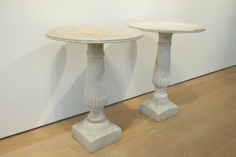 A pair of 19th century Italian Carrara marble garden tables with circular tops on baluster column pediments with square plinths. Elegant gadrooning on the central column. The lower collar of one of the bases has been replaced. Fabulous Patina.