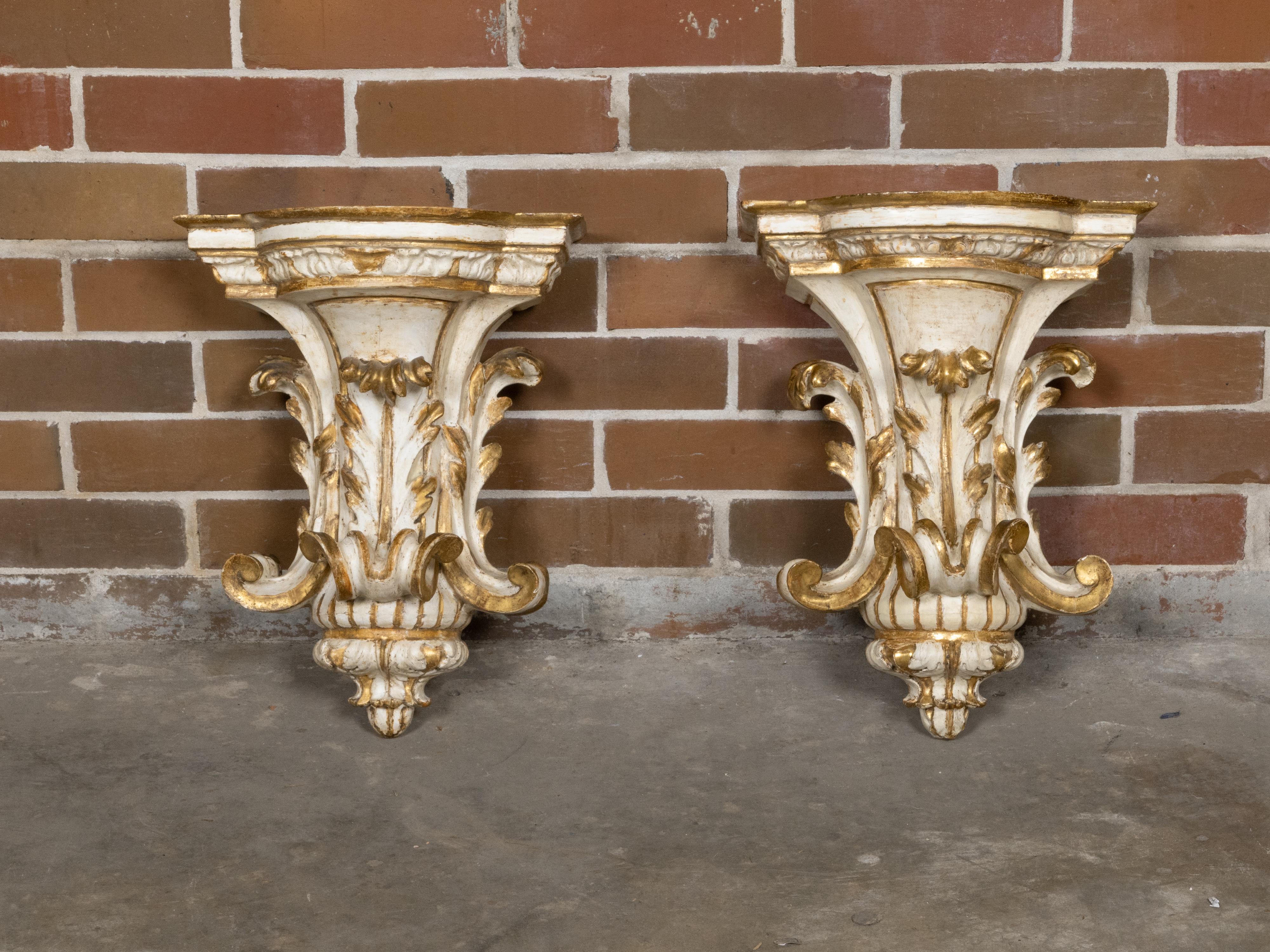 An ornate pair of 19th-century Italian wall brackets, hand-carved in wood and finished in a classic palette of ivory paint with gilded highlights. Each bracket is modeled as a corbel with a flared and stepped top, supported by exuberant acanthus