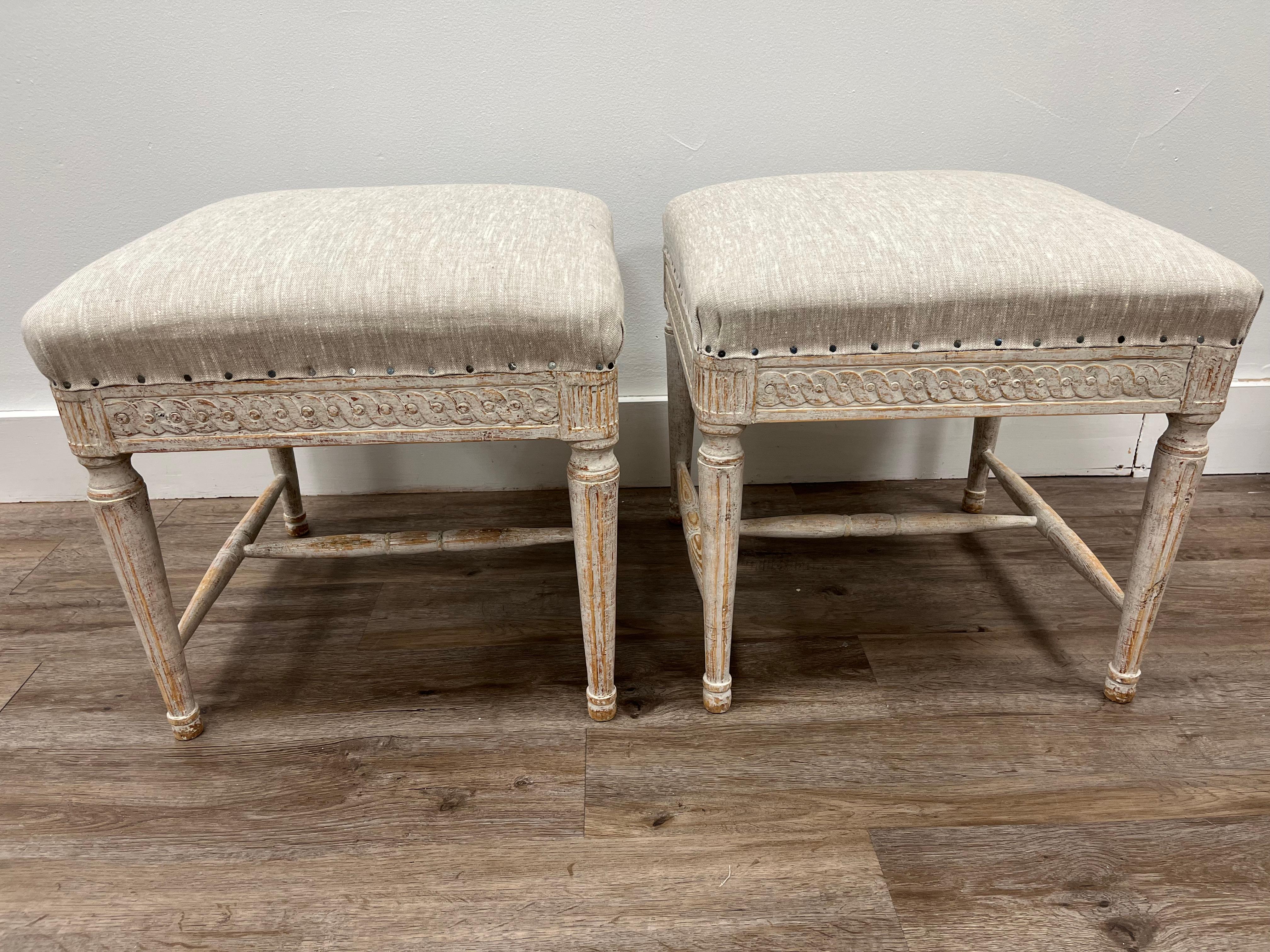A beautiful pair of Swedish Late Gustavian footstools. Featuring a decorative hand-carved Guilloché scroll with reeded fleurons on the corners. Turned legs are tapered and fluted. Supported by turned H-cross construction. Tastefully repainted in