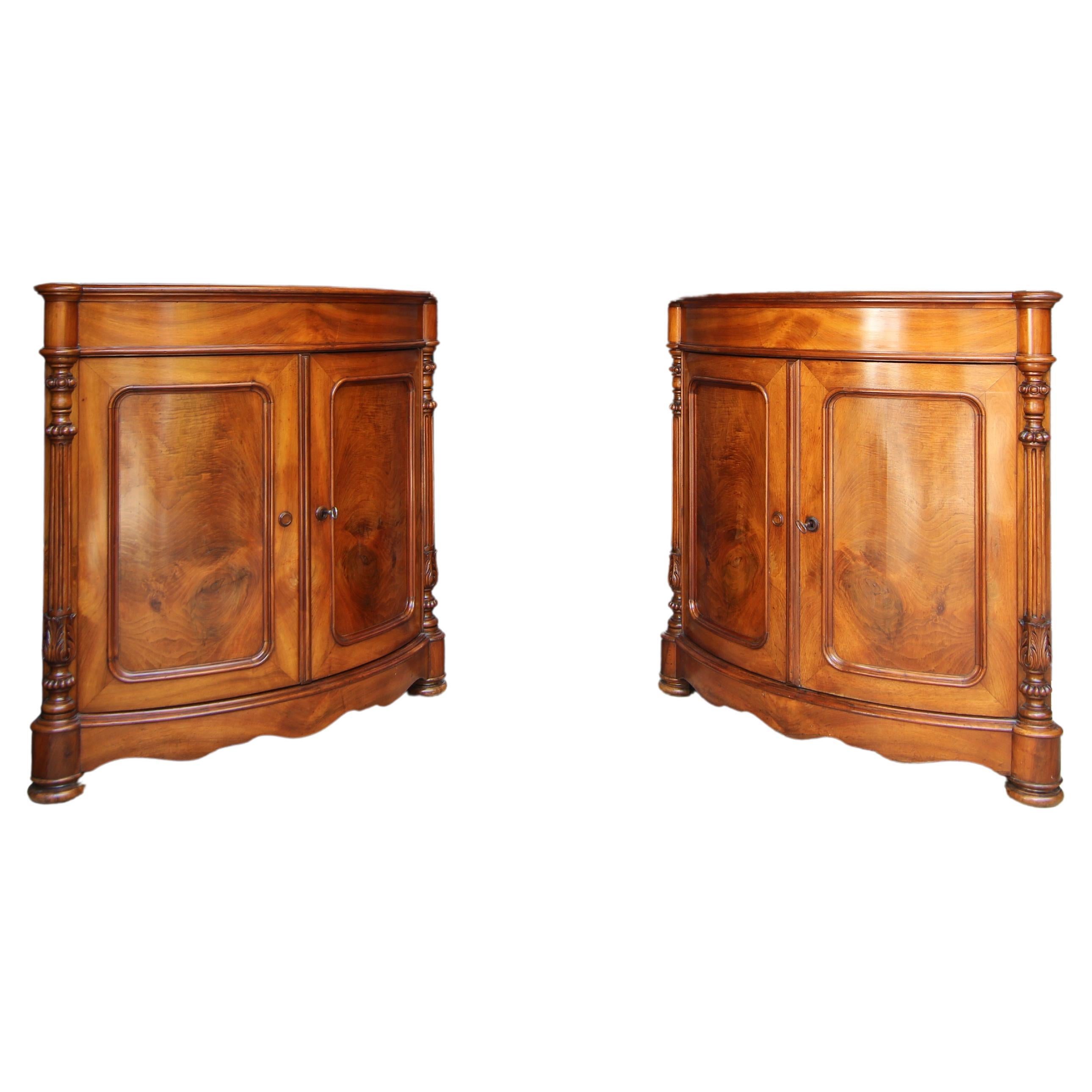 A pair of identical Louis Philippe corner cupboards from around 1860. Solid walnut and veneered on oak. 

Half-height two-door corner corpus with curved front, curved plinth apron and fluted profiled top. Round pilasters with carved and turned, as