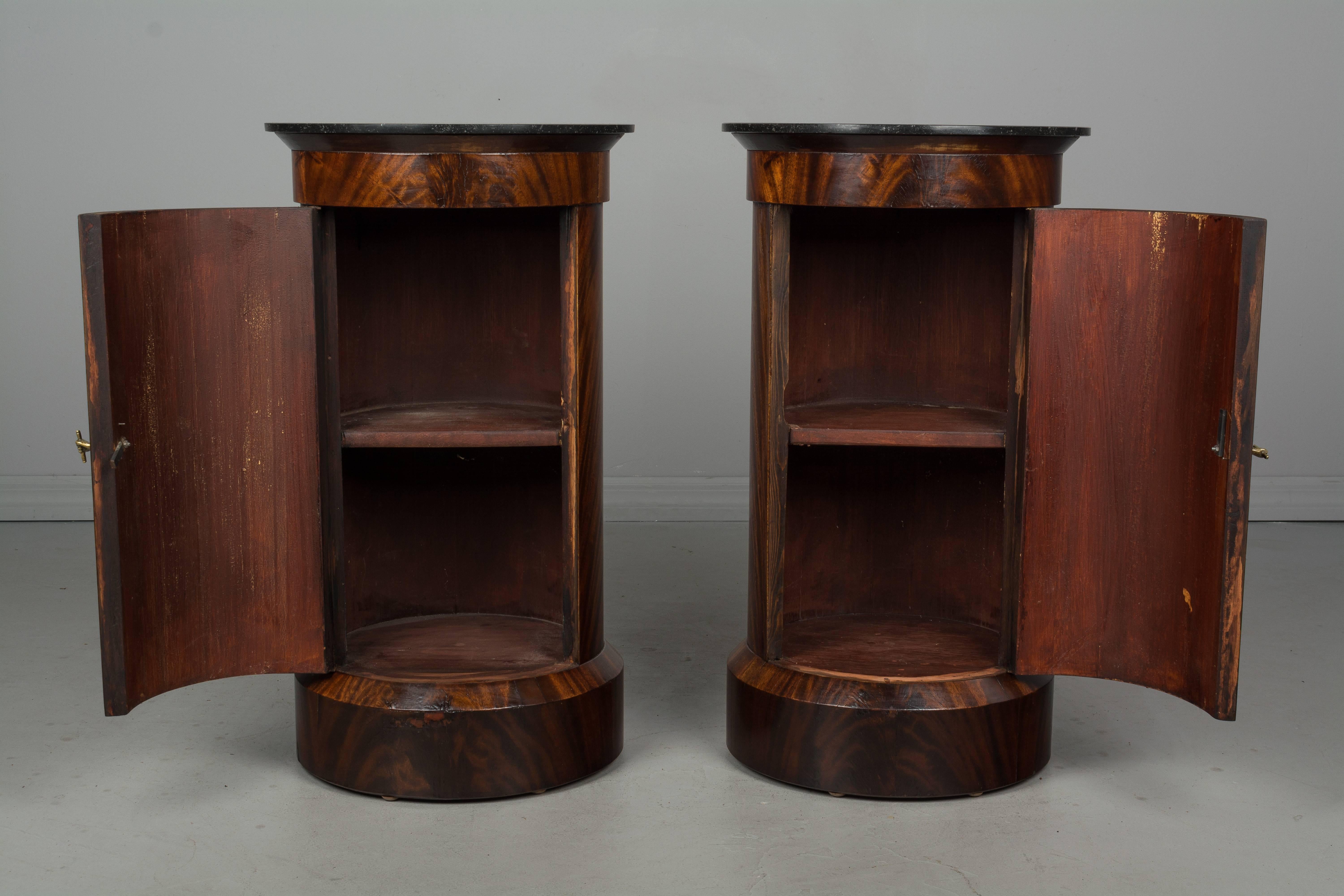 A pair of 19th century French Louis Philippe style mahogany pedestal side tables of cylindrical shape with dark grey marble tops. Single door with small brass hand knob opens to reveal a single shelf. Raised on a plinth base. Beautiful flame