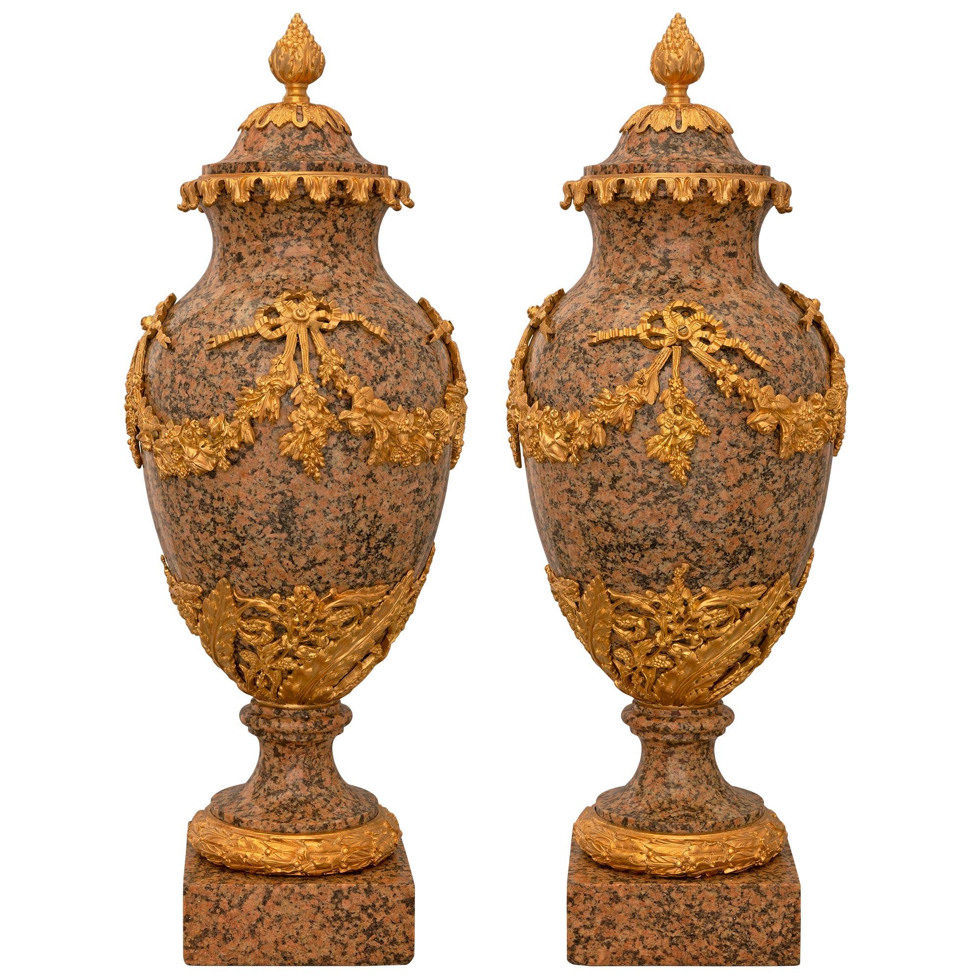 A spectacular and most impressive large scaled pair of 19th century French Louis XVI st. pink granite and ormolu mounted lidded urns. The urns are raised by a square base below a berried laurel ormolu band. The socles are below a richly chased
